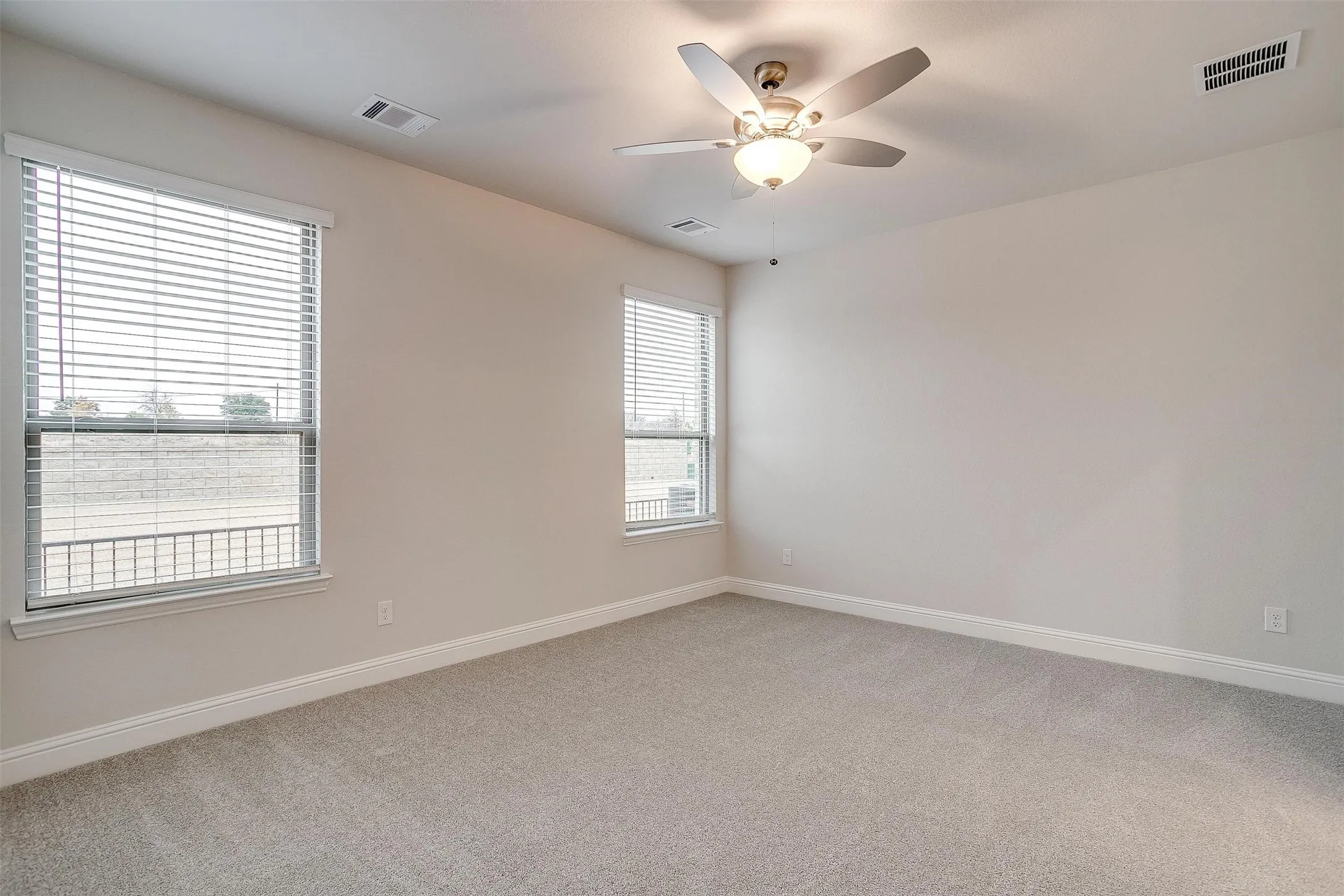 Neutral toned walls, light gray carpeting, two windows with blinds, and an overhead ceiling fan with light fixture