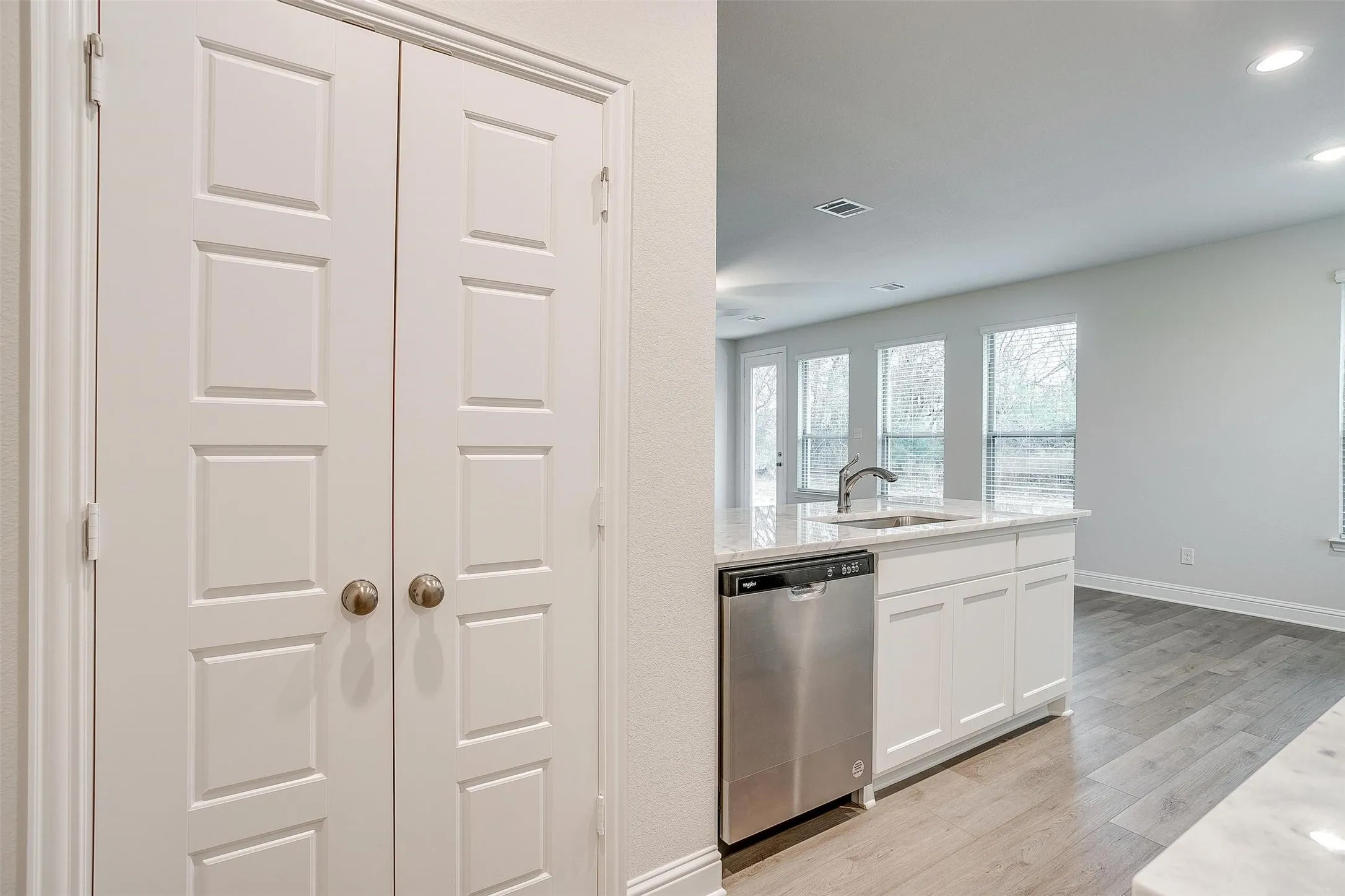 White paneled double doors, recessed lighting, white cabinetry, stainless steel dishwasher, and a kitchen island with a sink and faucet