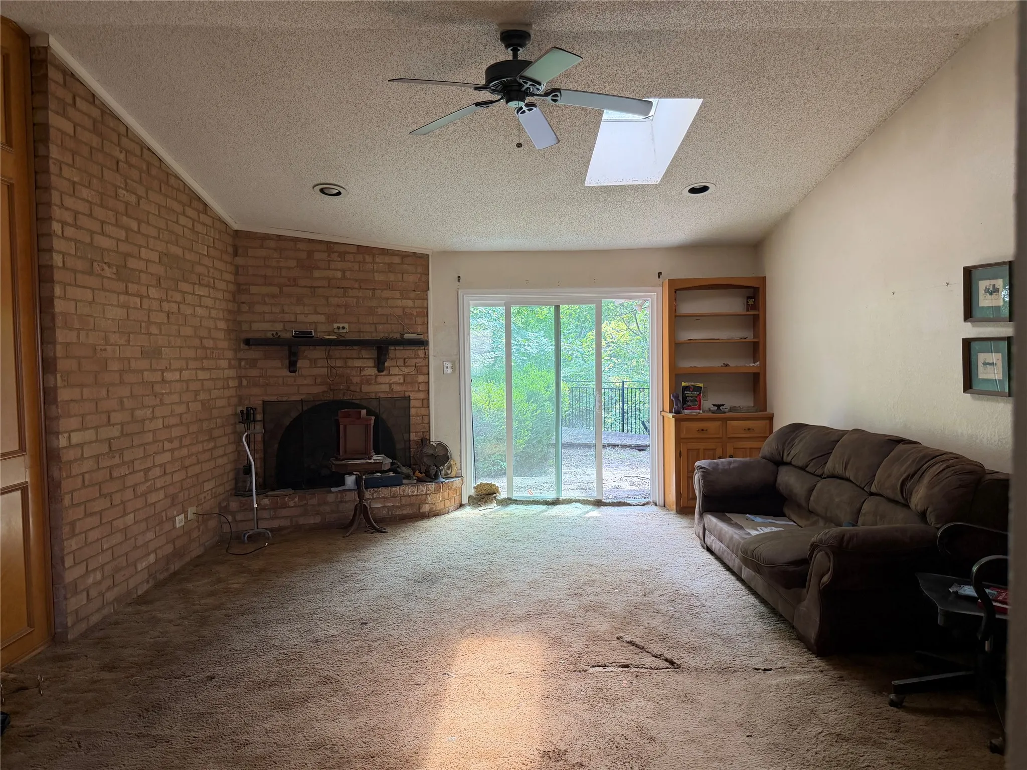 Brick-faced fireplace with hearth, ceiling fan, skylight, built-in shelving, and sliding glass door