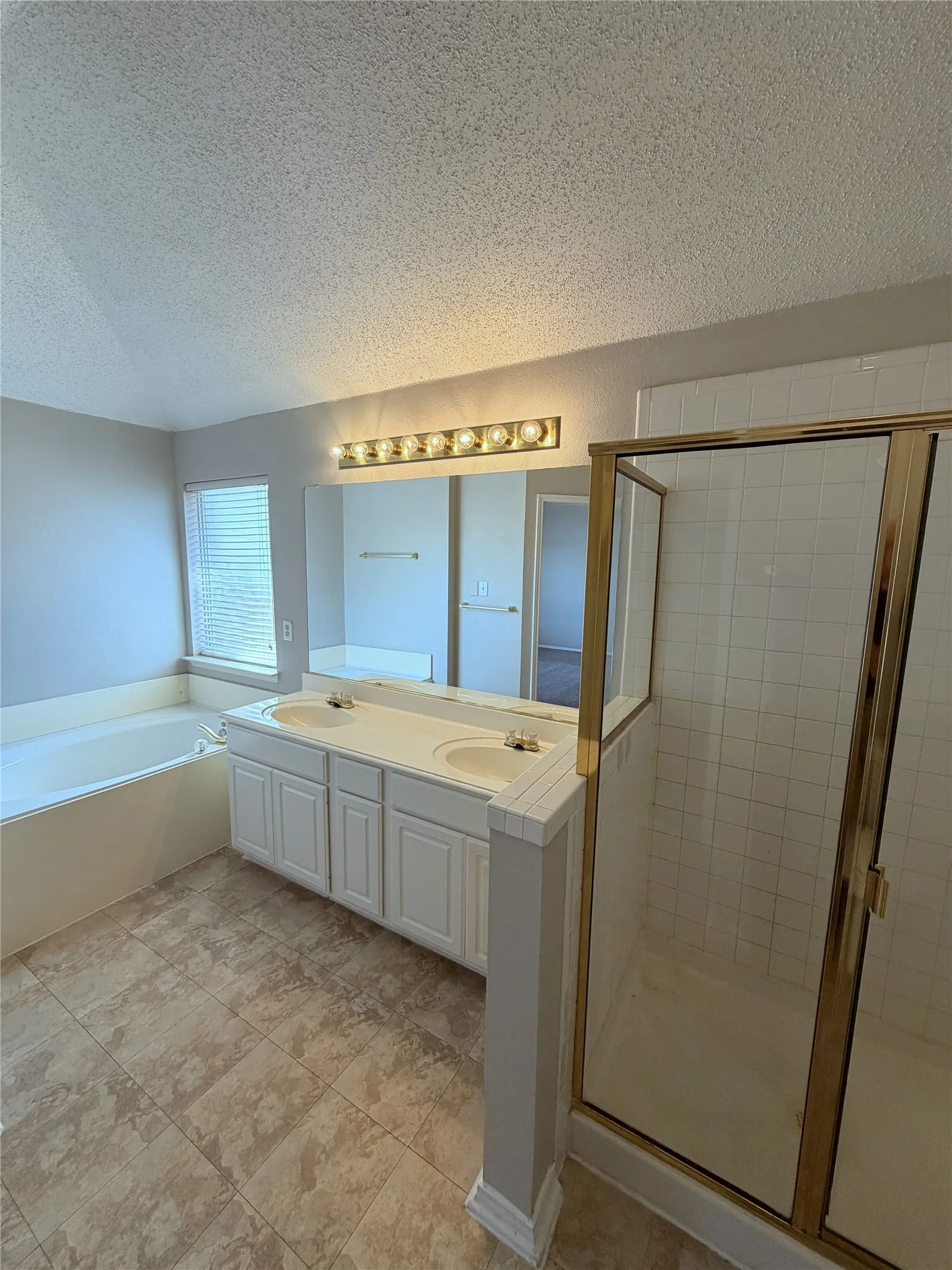 Dual vanity with white cabinetry, built-in soaking tub, tiled shower with glass enclosure, large wall-mounted mirror, and tiled flooring