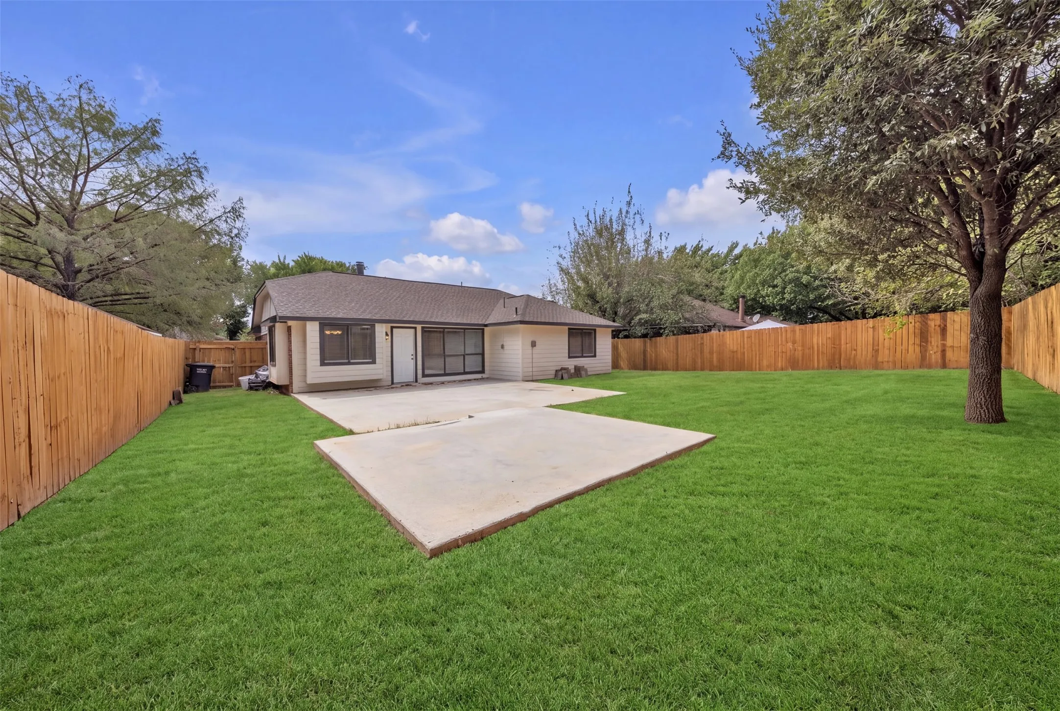 Expansive backyard featuring a manicured lawn, established shade tree, wood privacy fence, and a concrete patio. Grass is digitally enhanced