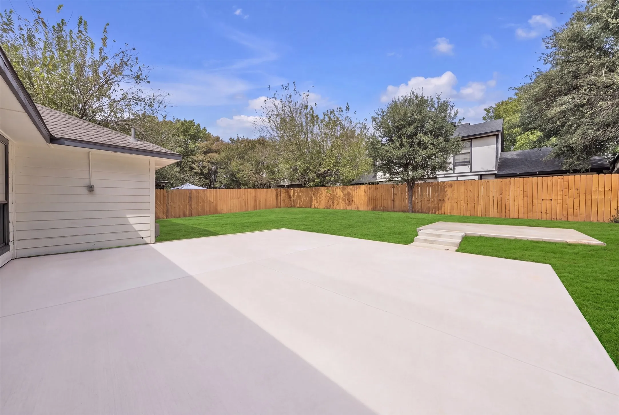 Expansive concrete patio with a step-up platform, verdant lawn, wood privacy fencing, and mature trees. Grass is digitally enhanced