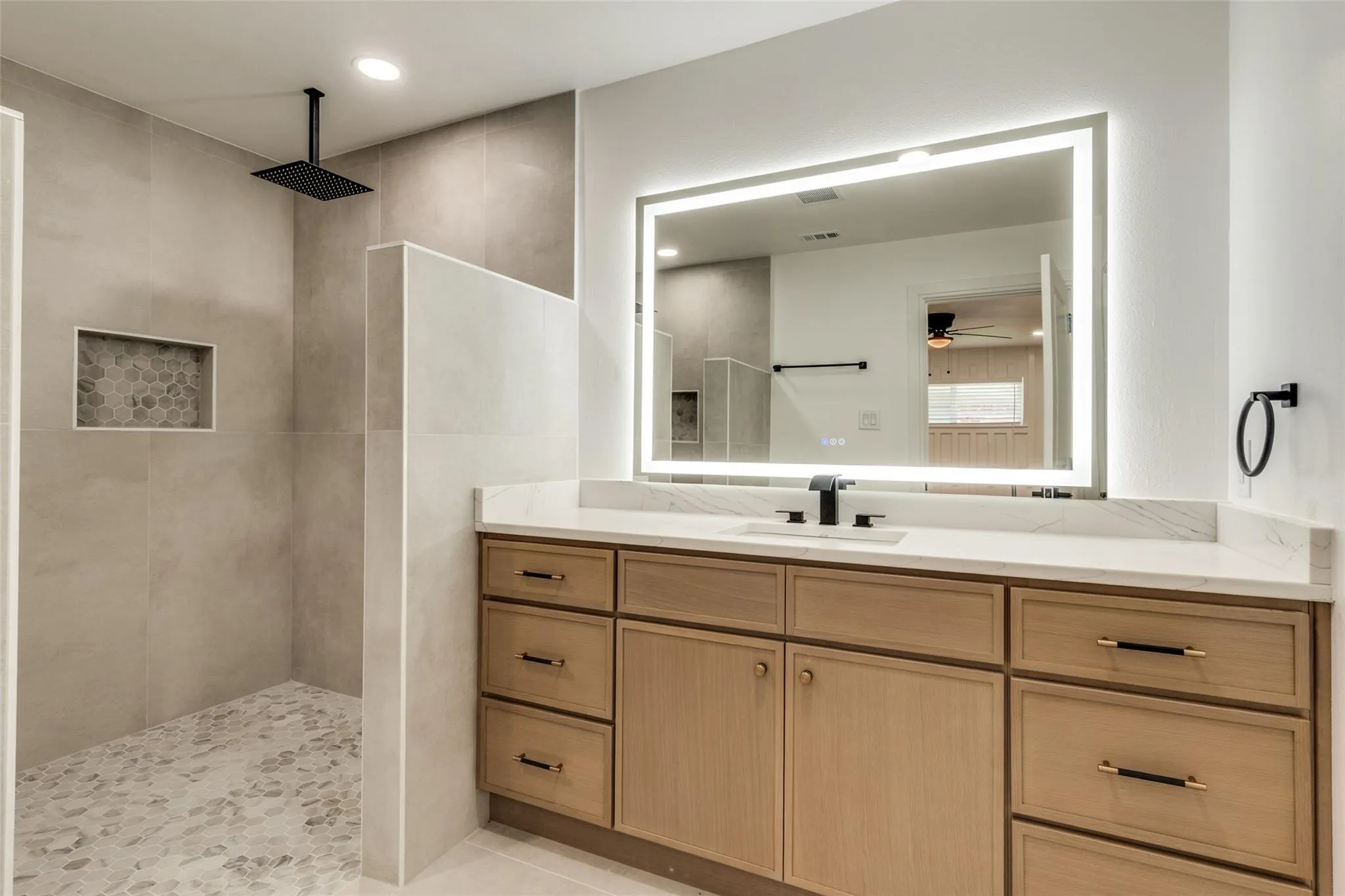 Walk-in shower with pebble flooring and recessed shelf, matte black rainfall showerhead, wood-finish vanity with stone countertop, matte black faucet, and illuminated mirror