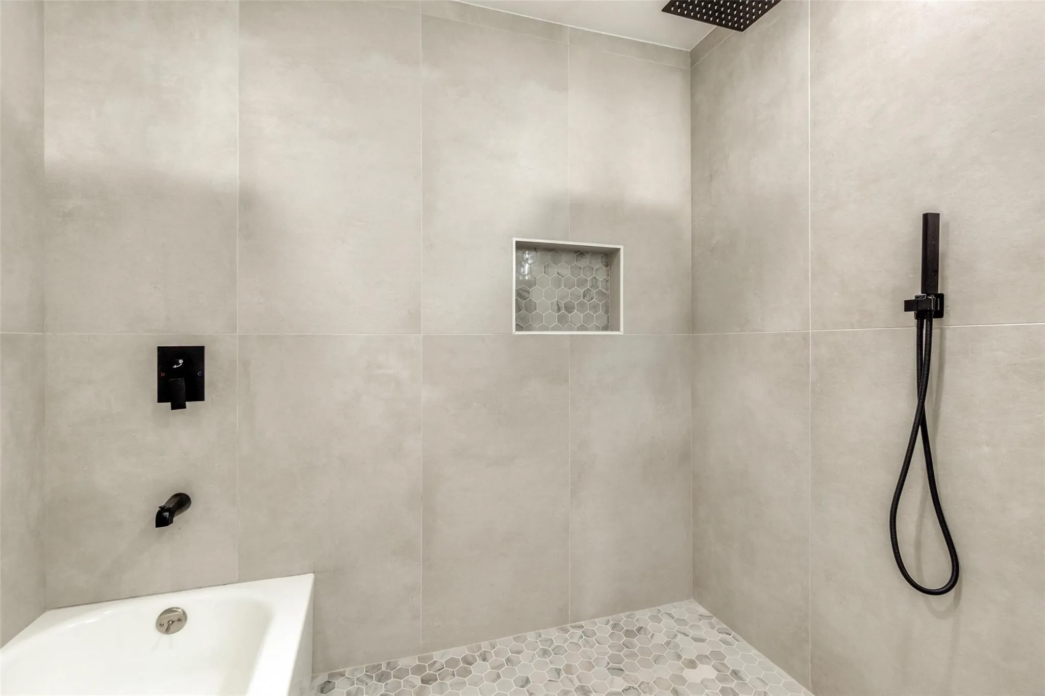 Bathtub and shower combination featuring matte black fixtures, an integrated shower niche with hexagonal mosaic tile, large format wall tiling, and a pebble tile shower floor
