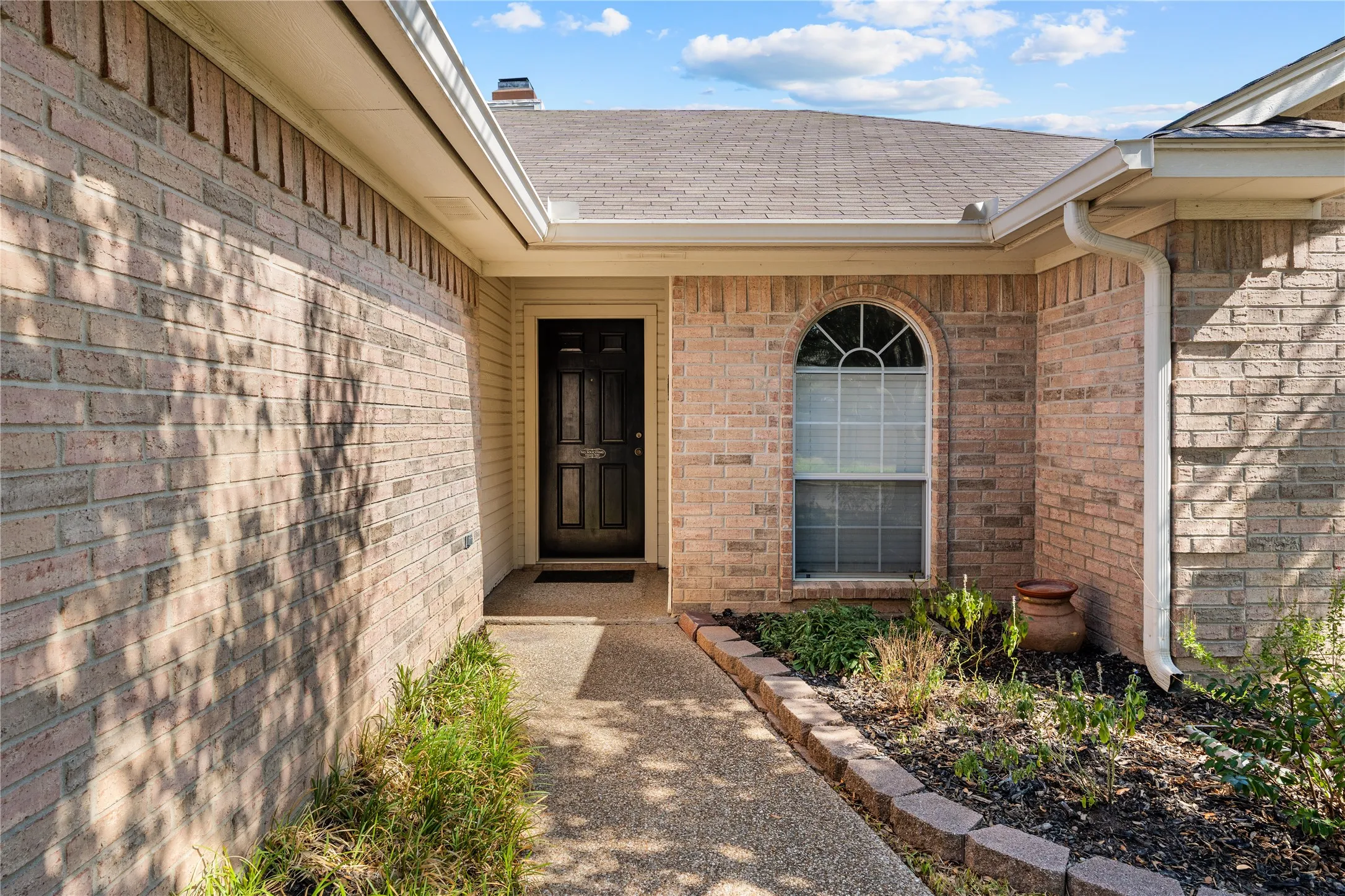 Brick exterior with an arched window, dark paneled entry door, concrete walkway, and landscaped garden bed with edging