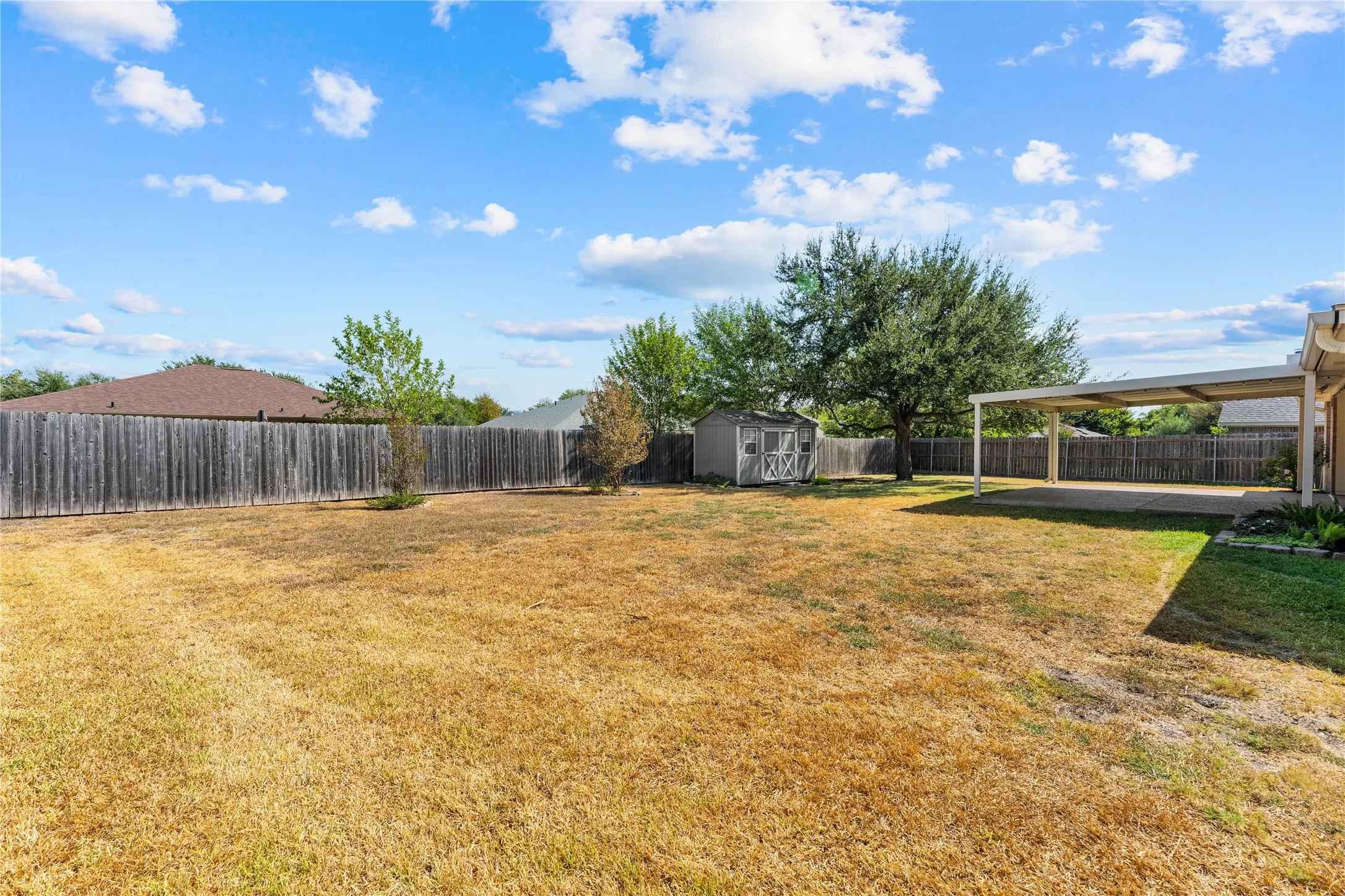 Expansive outdoor space featuring a covered patio, a detached storage shed, mature trees, and a perimeter wood fence