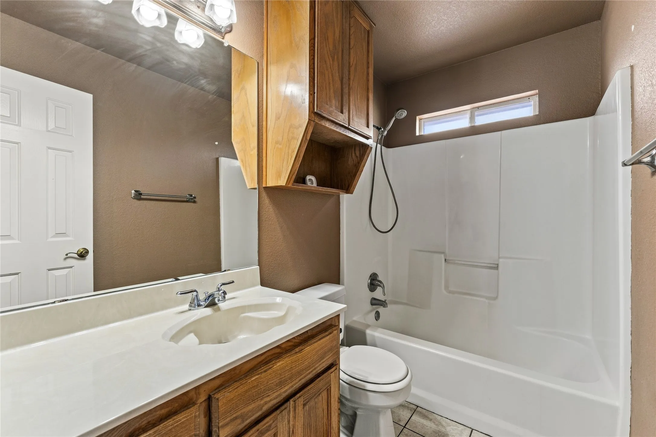 Vanity with integrated sink, wood-finish cabinetry, full-width mirror, shower/tub combination, and an upper window