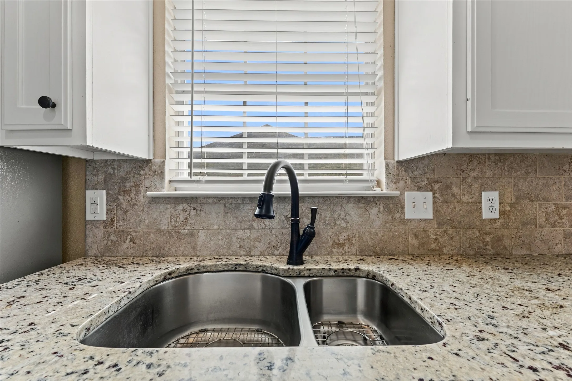 White shaker-style cabinetry with black hardware, stone-finish countertop, stainless steel double basin sink, matte black gooseneck faucet, and stone tile backsplash