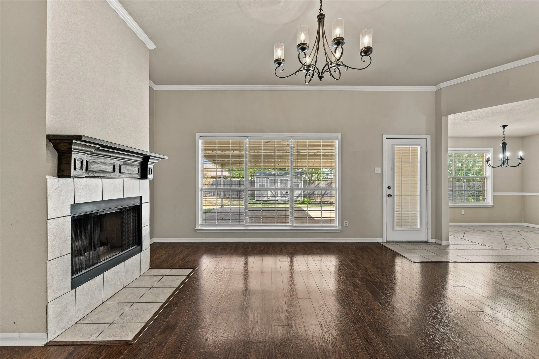 Wood-finish flooring, tiled fireplace with a dark wood mantel, large window with blinds, and a glass-paneled door