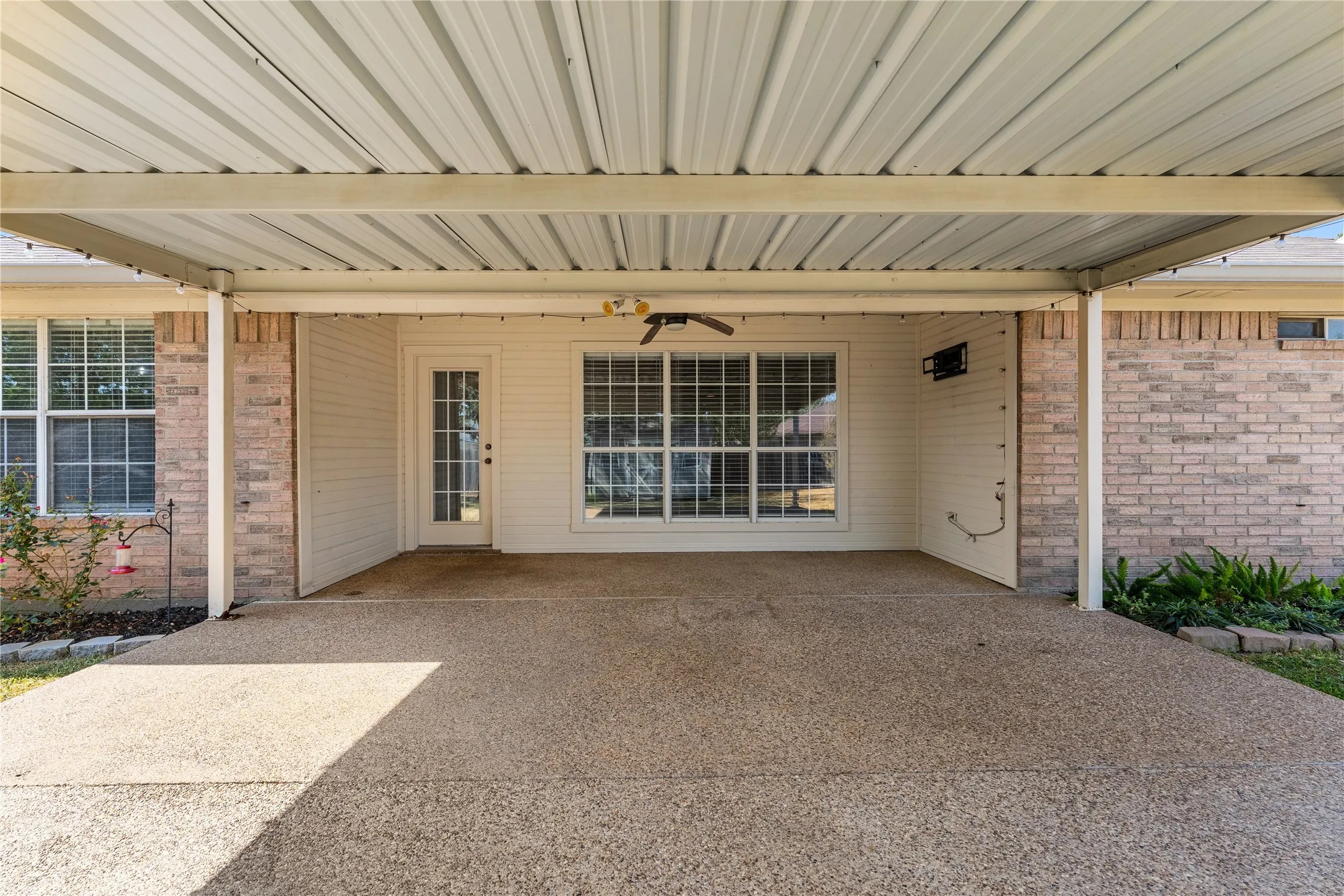 Expansive covered patio with exposed aggregate concrete flooring, featuring a ceiling fan and exterior lighting, alongside a brick and siding exterior