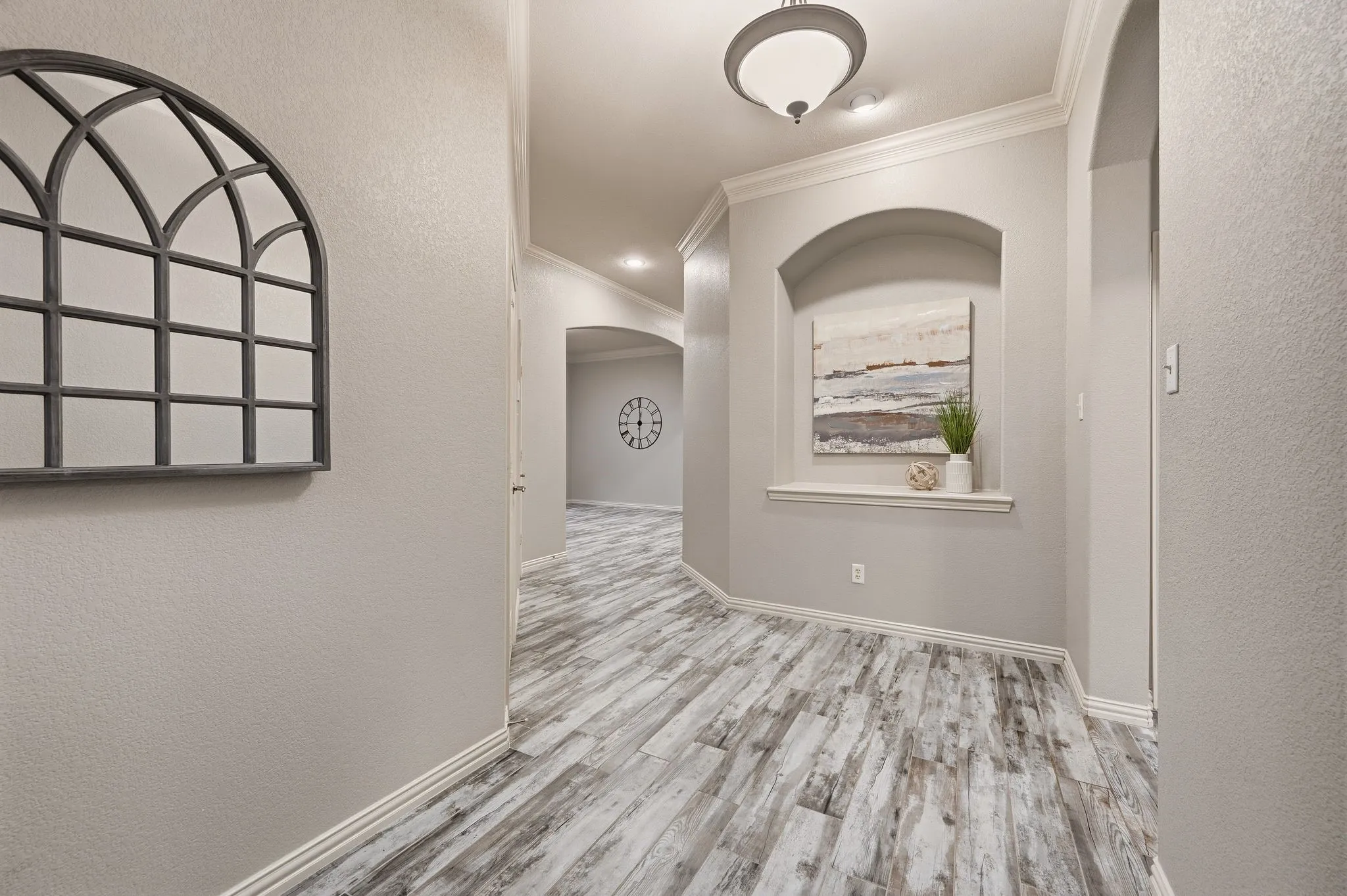 Elegant hallway showcasing wood-finish plank flooring with gray and white tonal variation, arched architectural alcove with decorative window treatment, recessed ceiling lighting, and geometric wall mirror accent