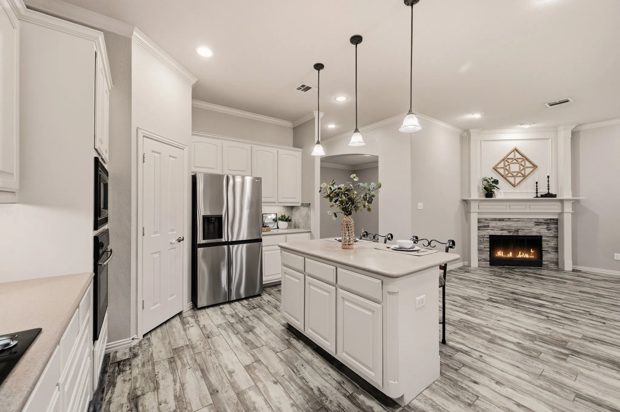 Kitchen island with pendant lighting, stainless steel appliances, wood-finish flooring, white cabinetry, and a fireplace with a stone surround