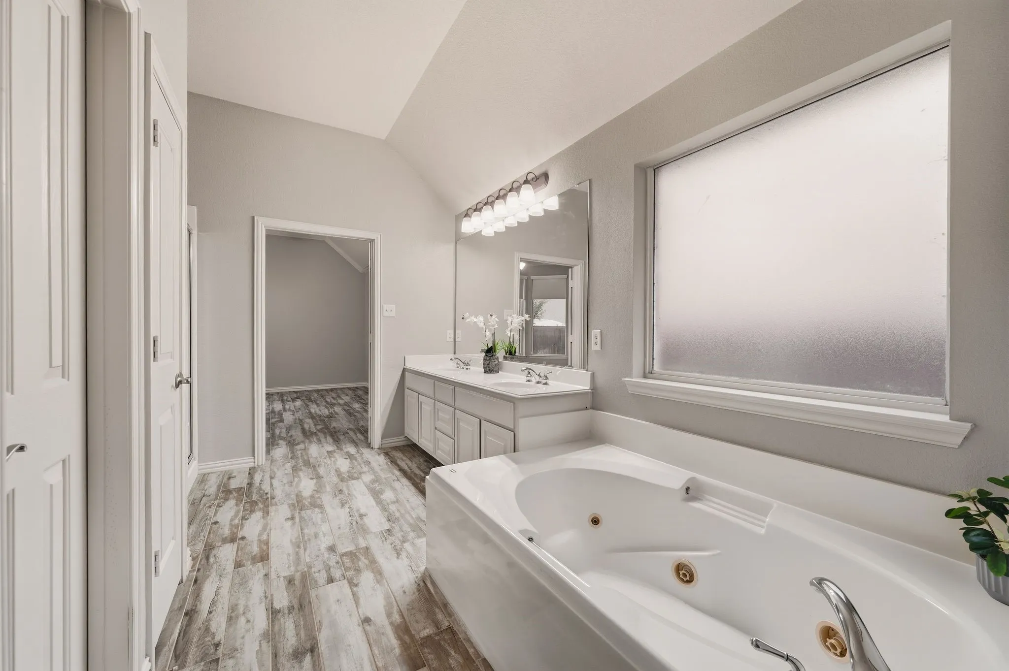 Vaulted ceiling, dual vanity with undermount sinks, wood-finish flooring, white soaking tub with jets, and frosted window