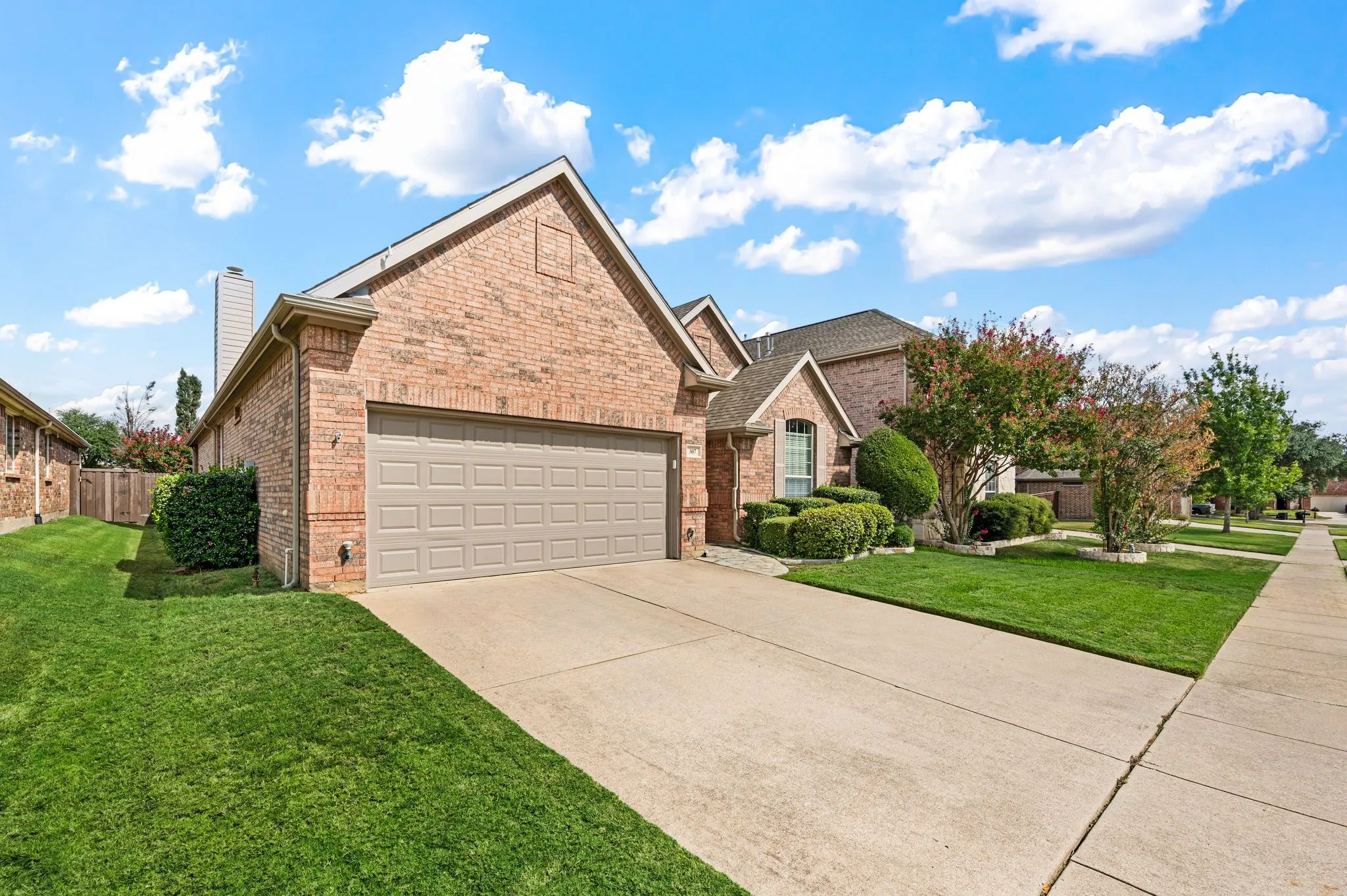 Brick exterior with an attached garage, concrete driveway, and established front landscaping