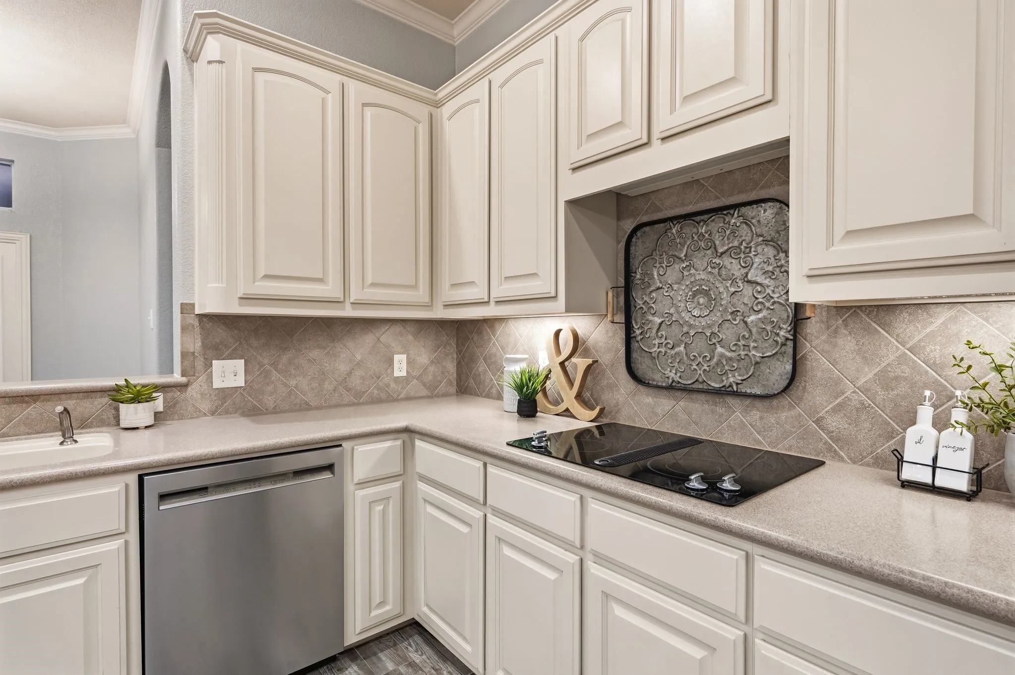 Built-in stainless steel dishwasher, integrated cooktop, light-toned cabinetry with panel detailing, light-toned countertops, and diagonal tile backsplash