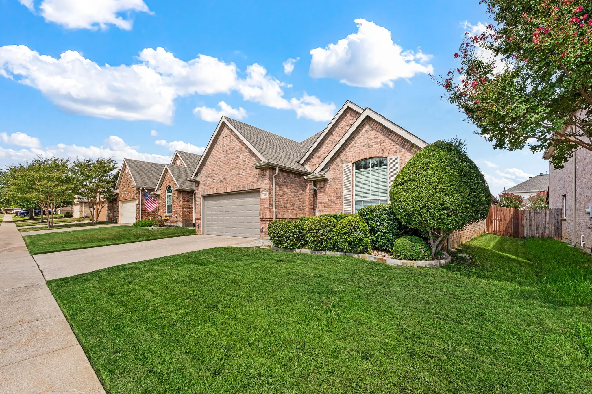 Brick exterior with shutters, an arched window, an attached garage, and a manicured lawn with mature landscaping