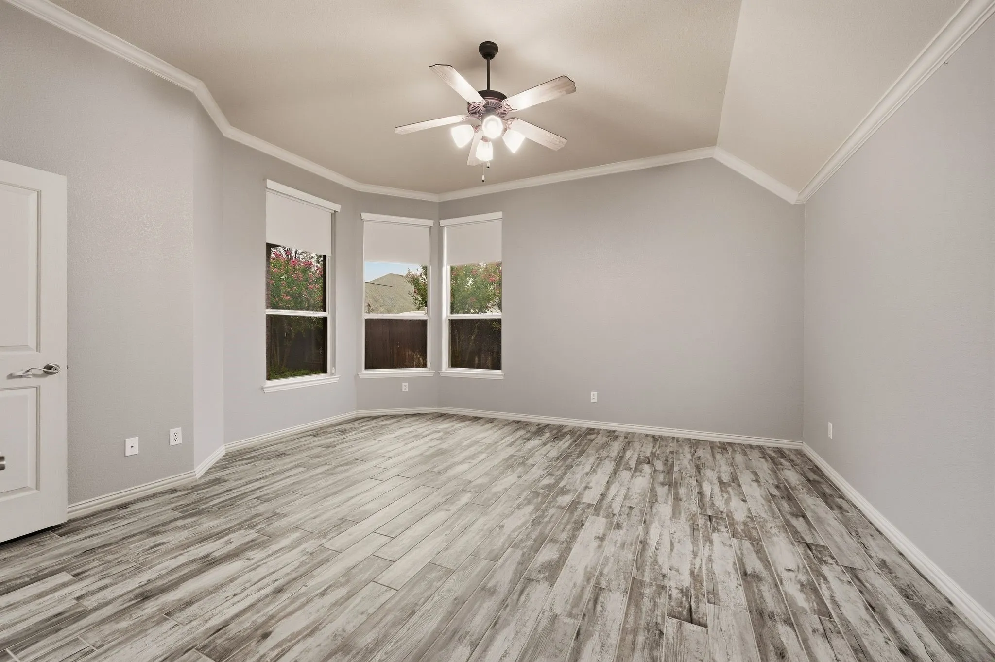 Vaulted ceiling with crown molding, wood-finish flooring, ceiling fan with integrated lighting, and a bay window featuring roller blinds