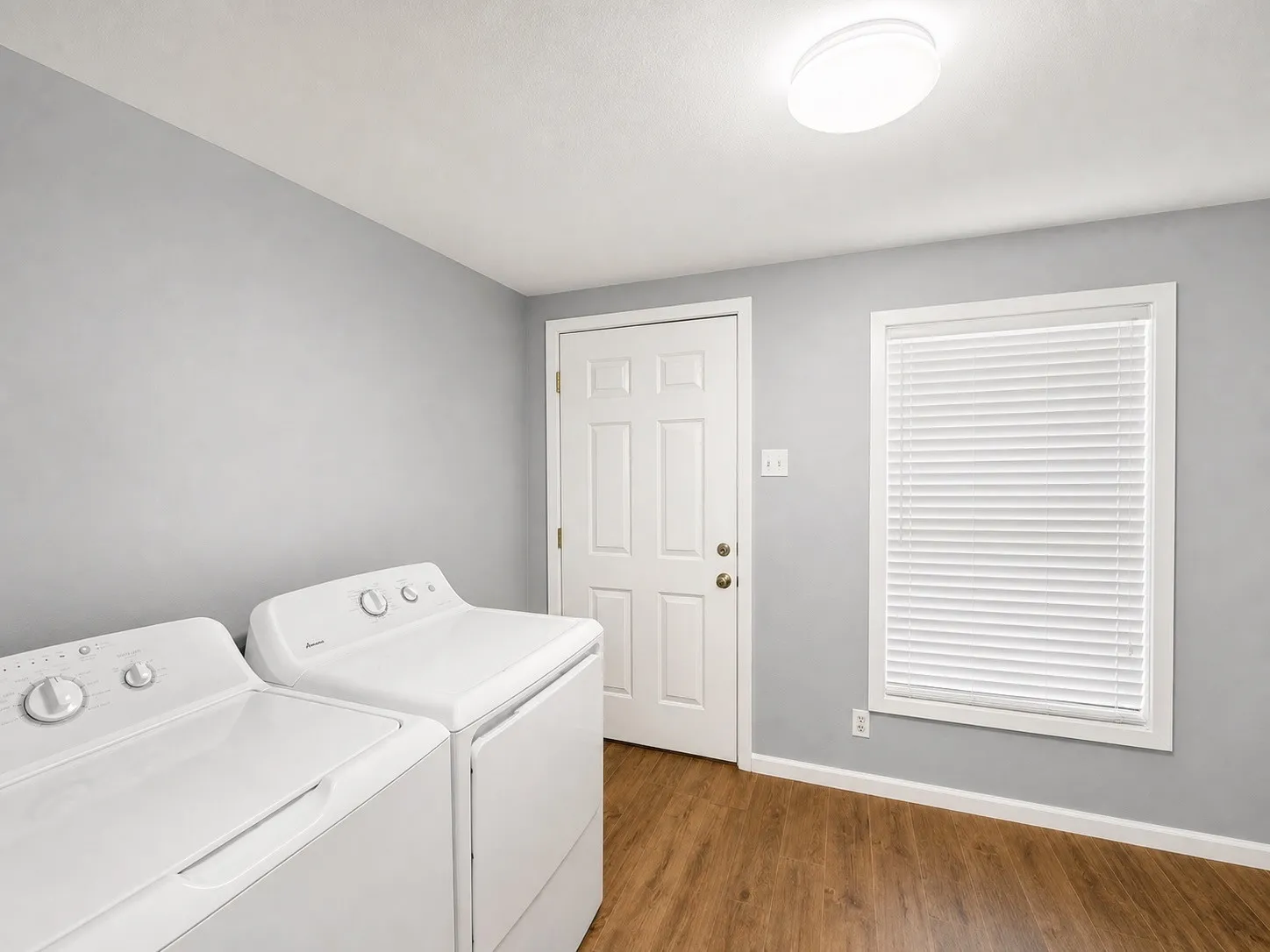 Functional laundry area featuring wood-finish flooring, a ceiling-mounted light fixture, and a window with blinds