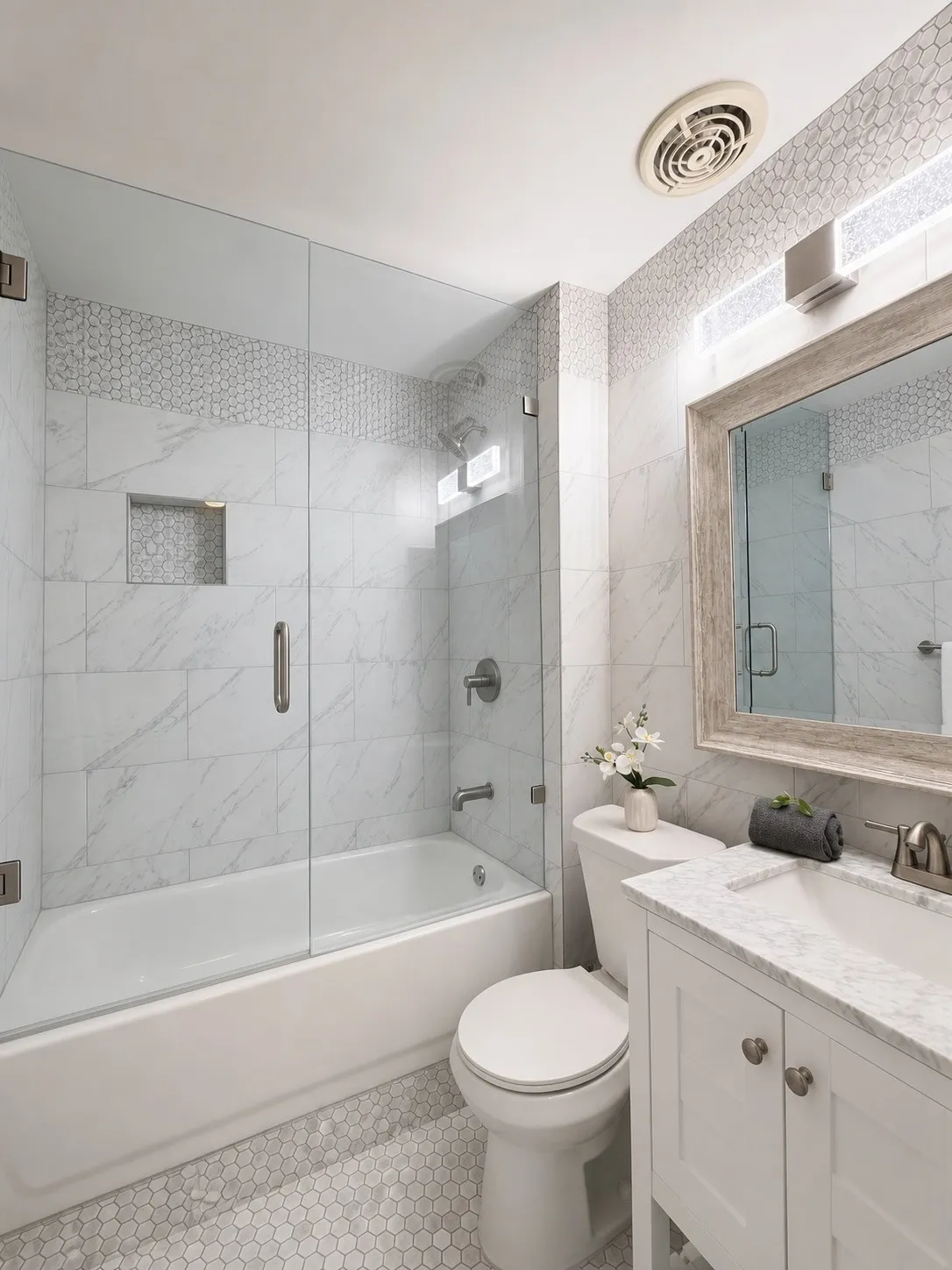 Bathroom featuring a white bathtub with a clear glass enclosure, marble-look wall tiling with decorative accent, hexagon mosaic floor tiles, a white vanity with a marble-look countertop, and a framed mirror with integrated lighting