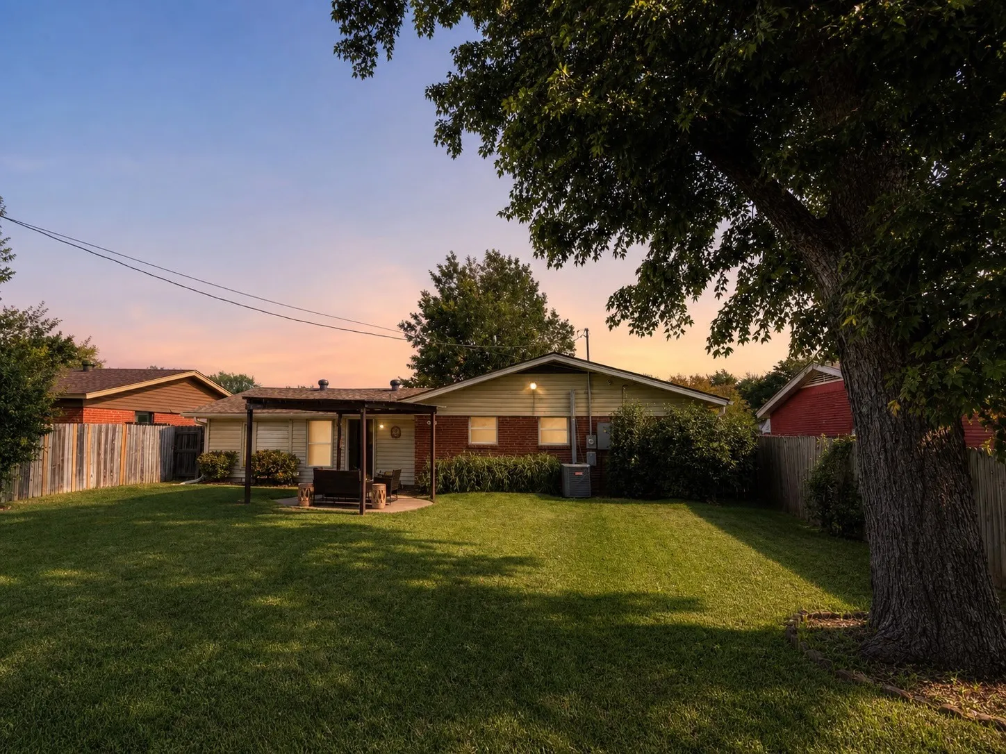 Expansive grassy yard with a wooden privacy fence, mature trees, a covered patio area, and a brick and siding exterior