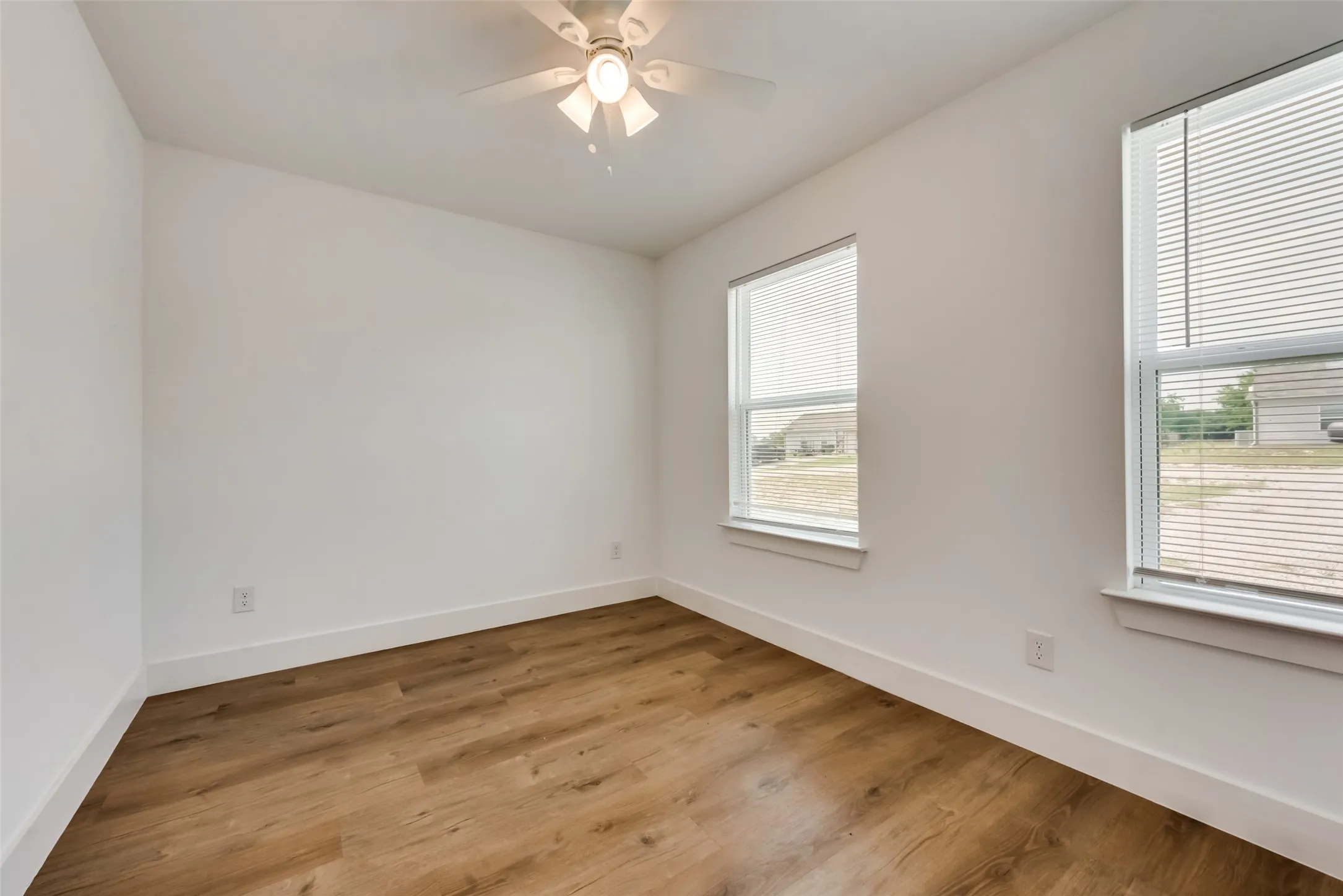 Neutral white wall finishes, wood-finish flooring, two windows with blinds, and a ceiling fan with integrated lighting