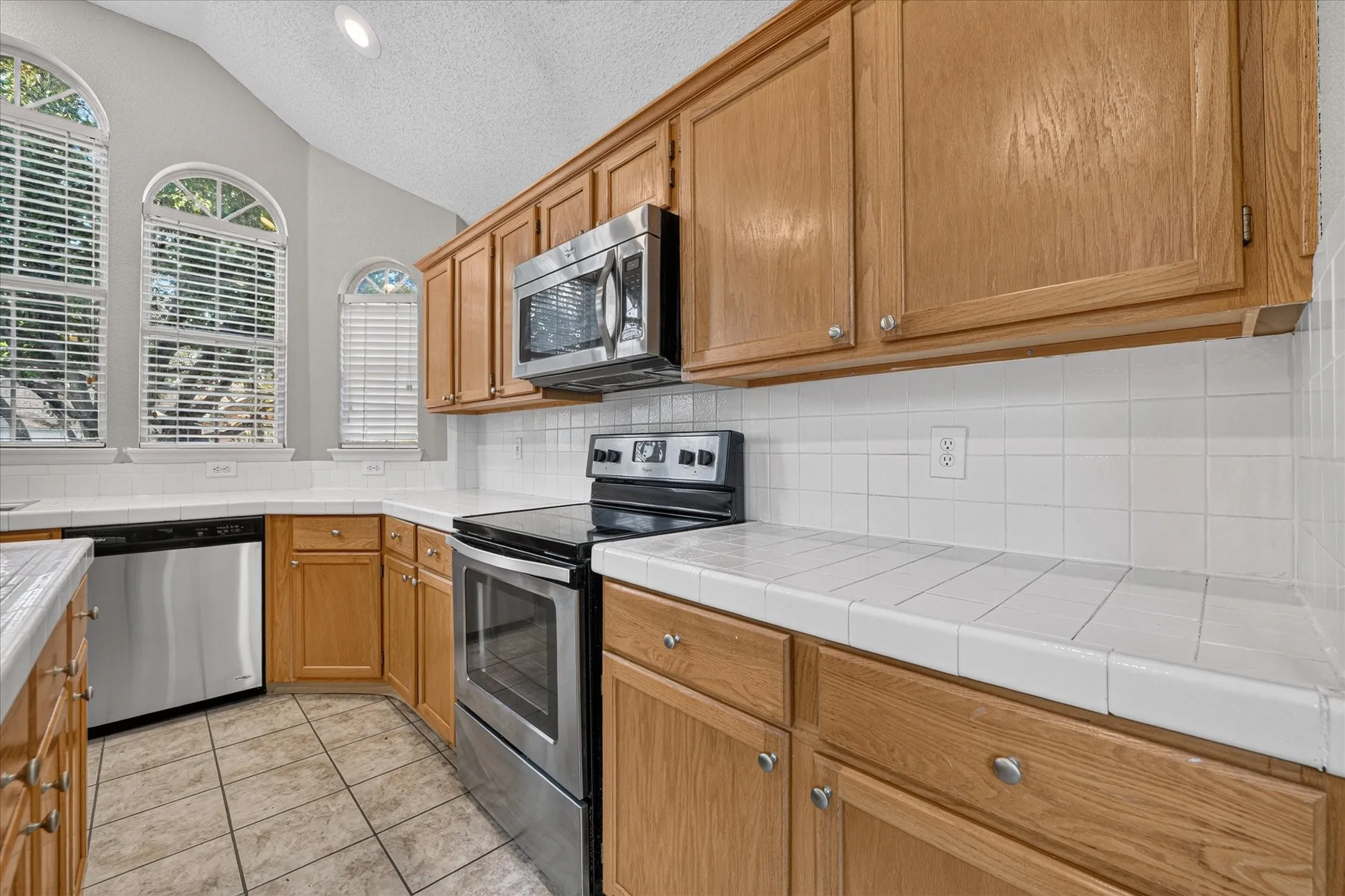 Kitchen with wood-finish cabinetry, tile countertops, stainless steel appliances, and arched windows