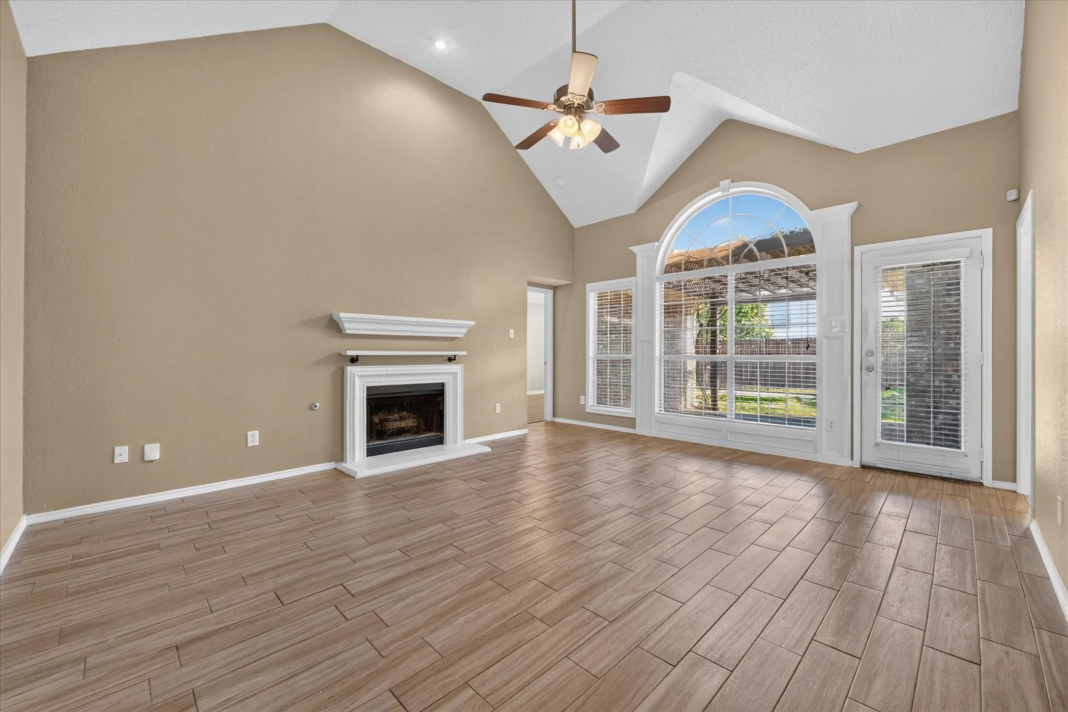 Vaulted ceiling with a ceiling fan, fireplace with a white mantel, wood-finish tile flooring, arched window with decorative trim, and an exterior door with integrated blinds