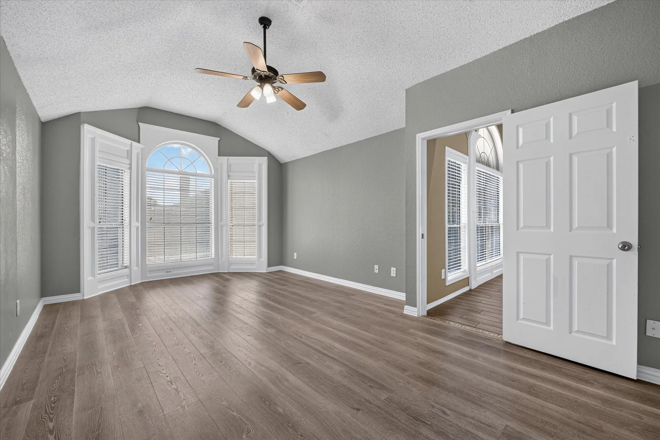 Vaulted ceiling with a ceiling fan, wood-finish flooring, and a bay window featuring a large arched central pane