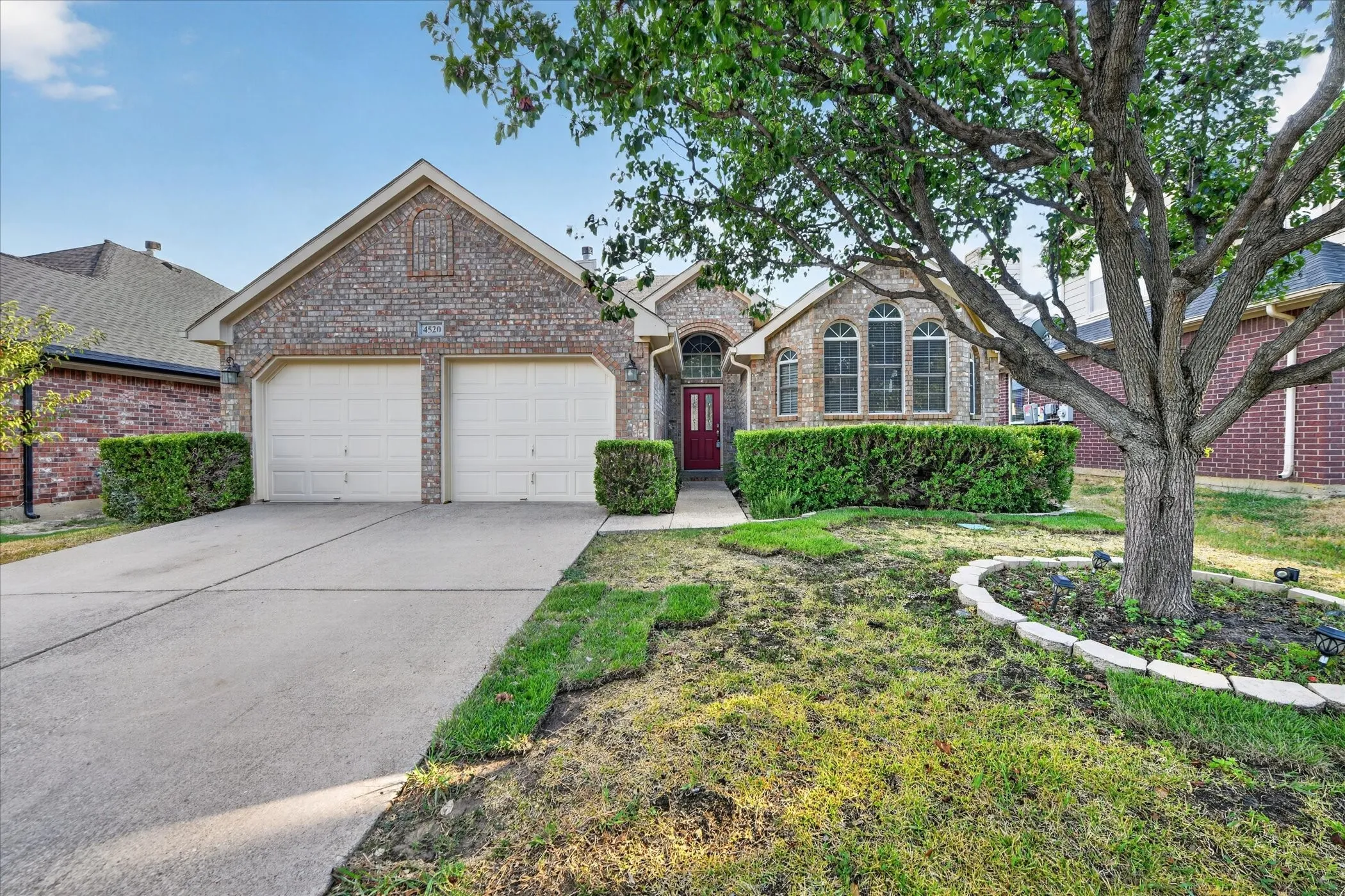 Brick exterior with arched window details, a prominent red entry door, a two-car garage, mature front yard tree, and established landscaping
