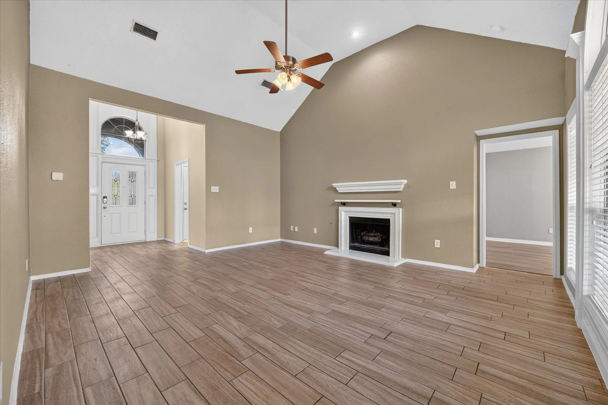 Vaulted ceiling with a ceiling fan, a decorative fireplace with a white mantel, wood-finish flooring, a white paneled entry door with sidelights, and vertical blinds