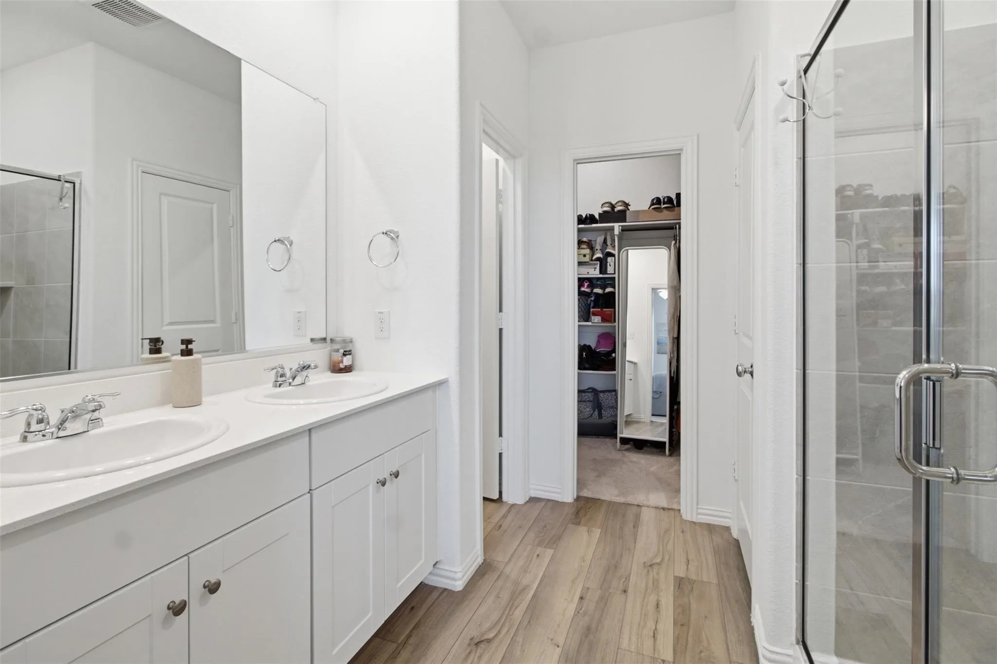Dual vanity with white cabinetry, wood-finish flooring, and a glass-enclosed shower