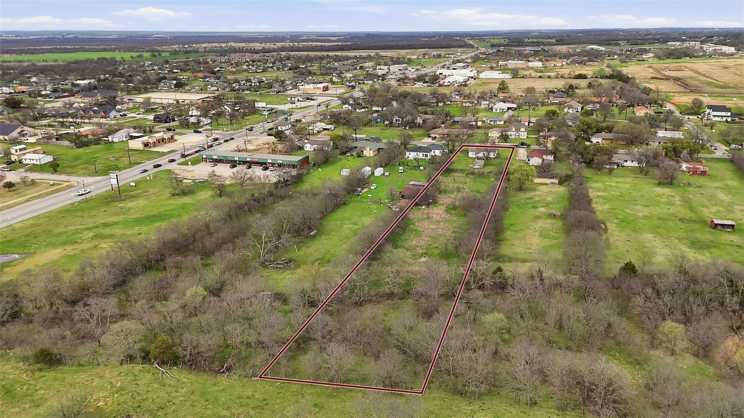 Expansive land parcel featuring a rectangular shape, bordering a wooded area, with scattered residential structures nearby