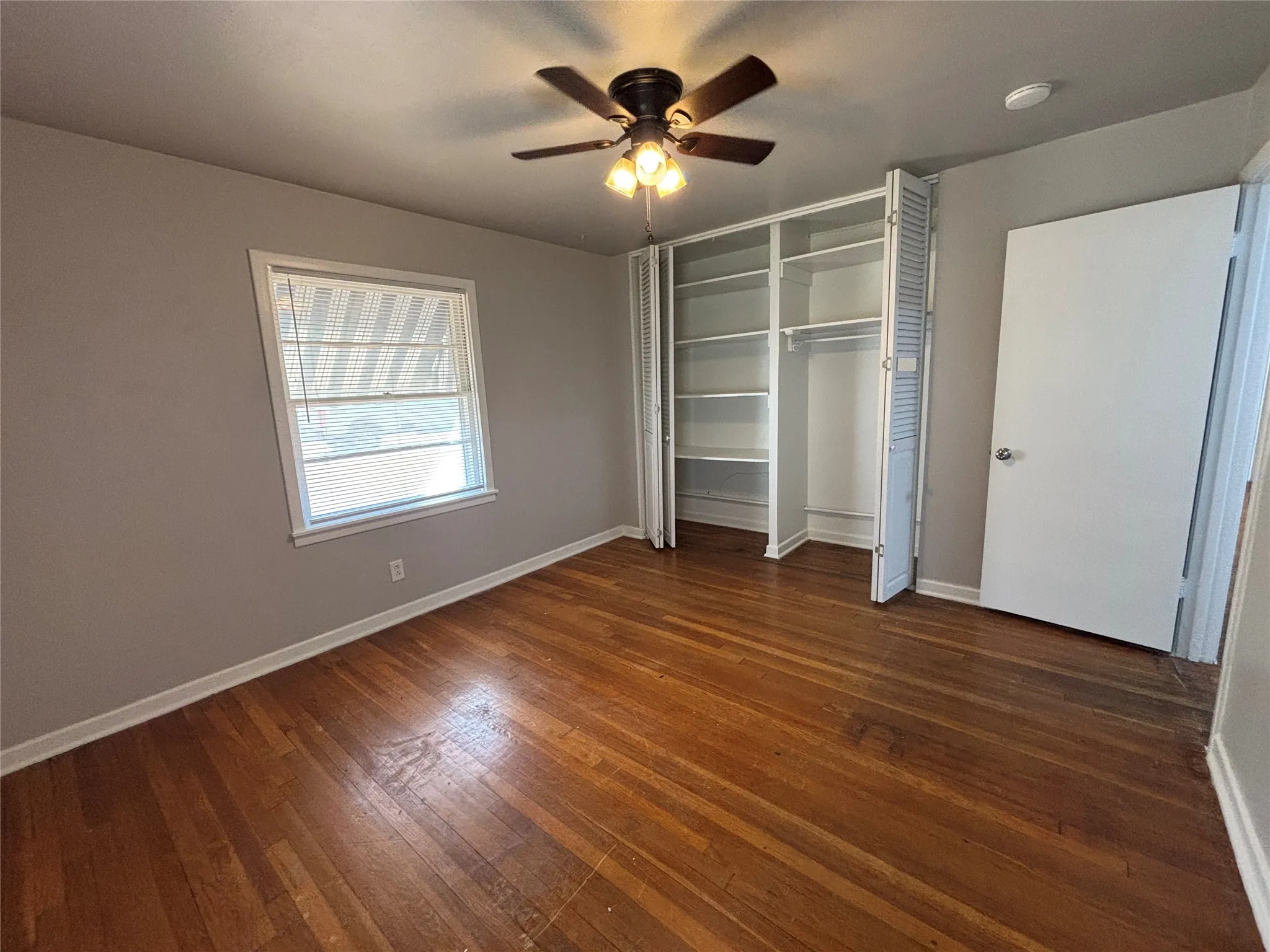 Wood-finish flooring, a window with blinds, a ceiling fan with integrated lighting, and built-in closet shelving