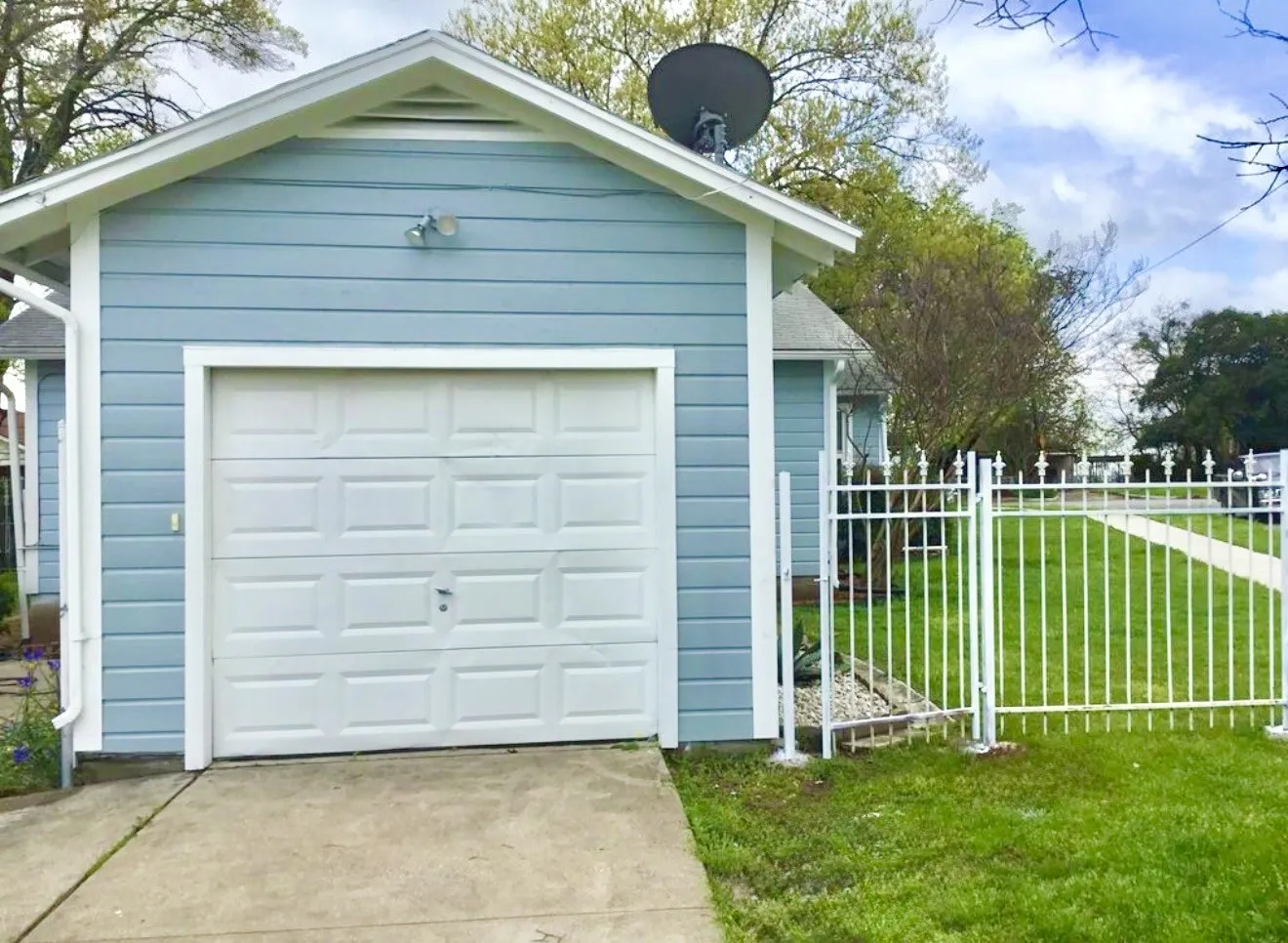 Detached garage with horizontal siding, a paneled garage door, concrete driveway and white metal fence.