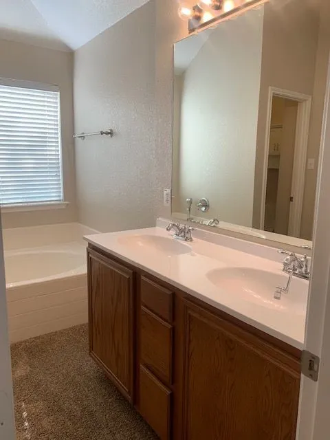 Dual sink vanity with wood-finish cabinetry, integrated countertop, and an oval soaking tub with window