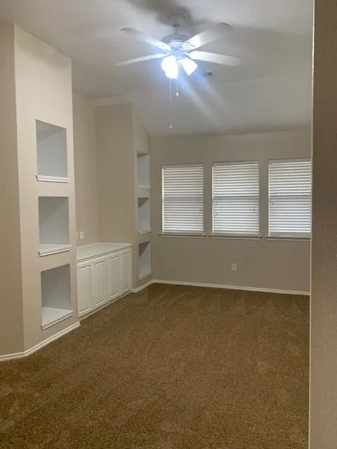 Carpeted room featuring built-in shelving, white cabinetry, multiple windows with blinds, and a ceiling fan with integrated lighting