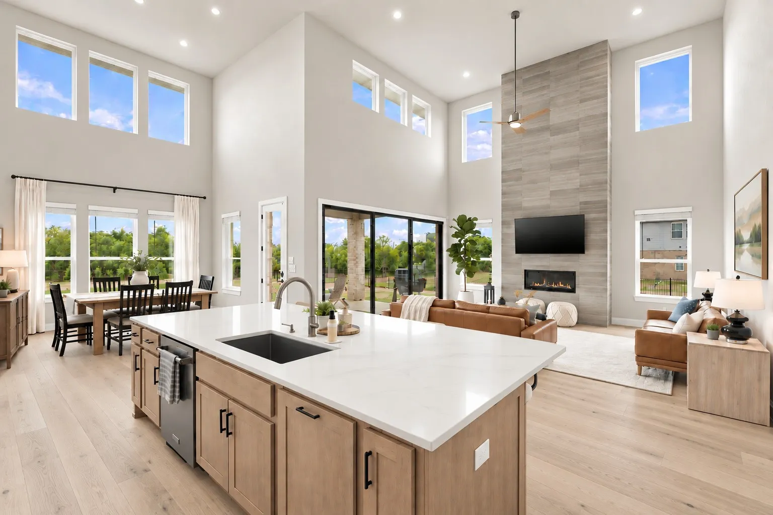 Open-concept interior featuring light wood-finish flooring, a kitchen island with a sink and white countertop, a dining area, a living area with a fireplace, and high ceilings with multiple windows