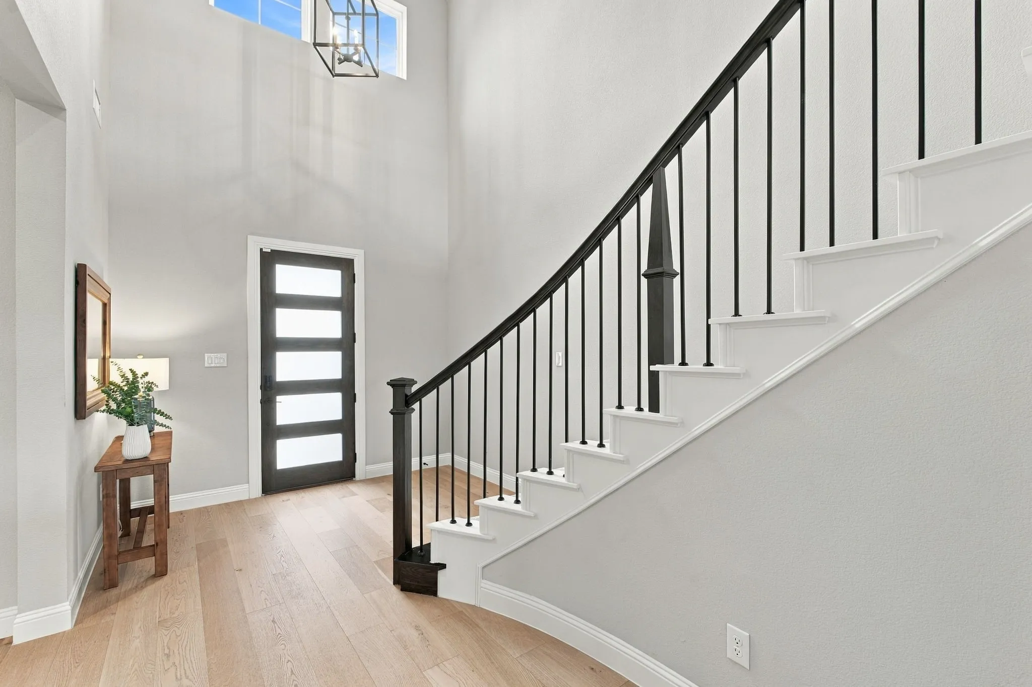 Two-story foyer with light wood-finish flooring, a modern front door with frosted glass panels, and a staircase featuring black metal balusters and a dark wood newel post