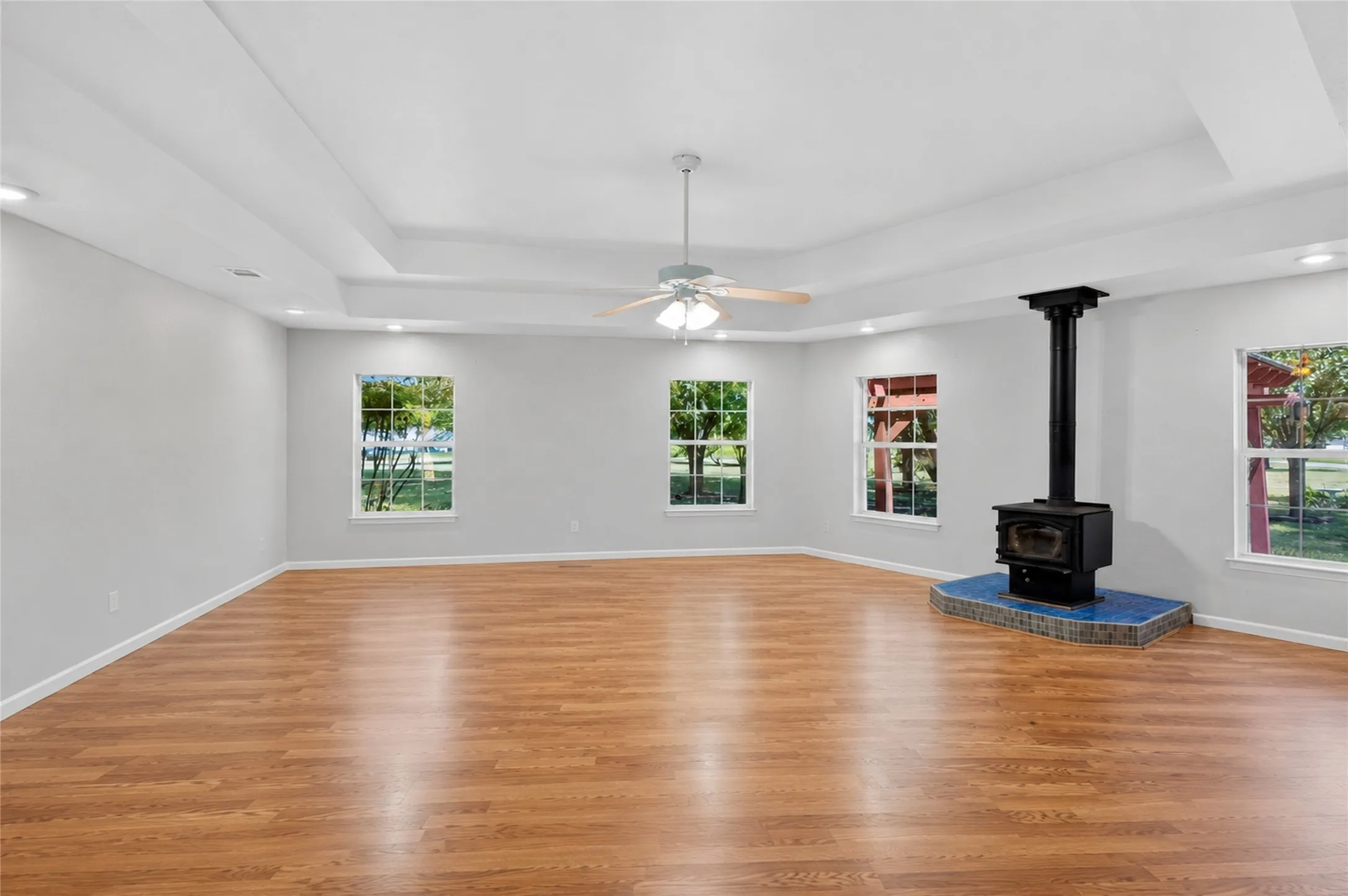 Expansive living area featuring wood-finish flooring, a wood-burning stove with a tiled hearth, tray ceilings, and recessed lighting - Virtually Cleared