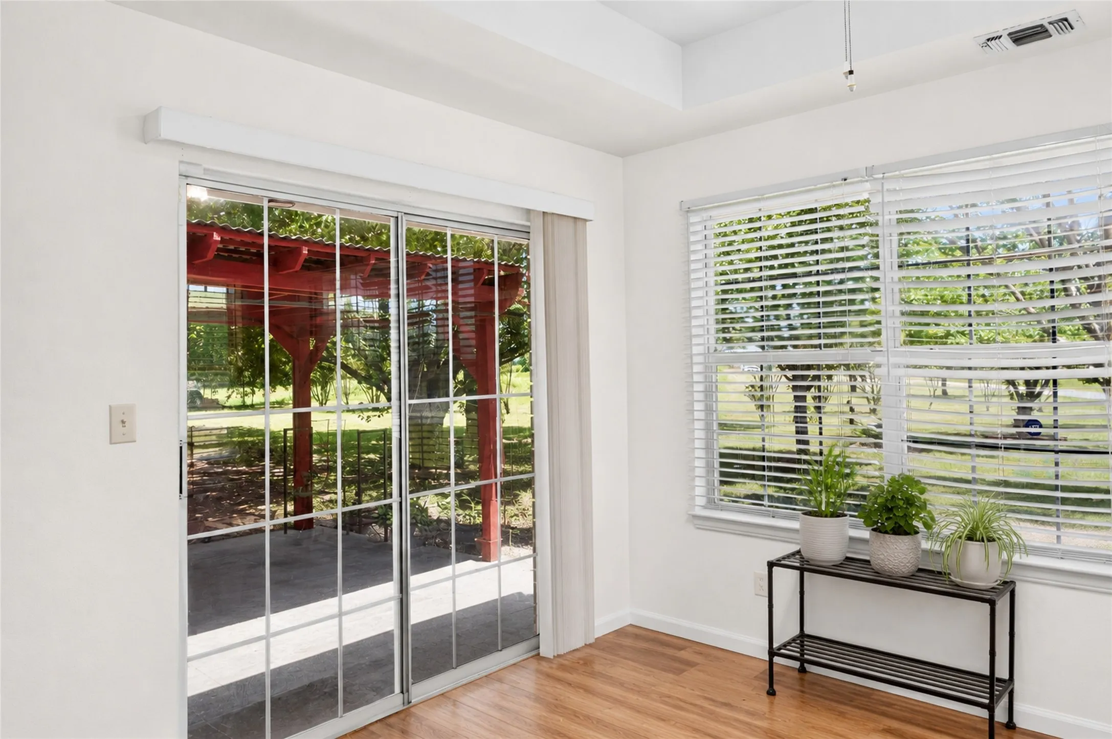 Sliding glass doors opening to an exterior patio with a red pergola, accompanied by a window with white blinds and wood-finish flooring - Virtually Staged