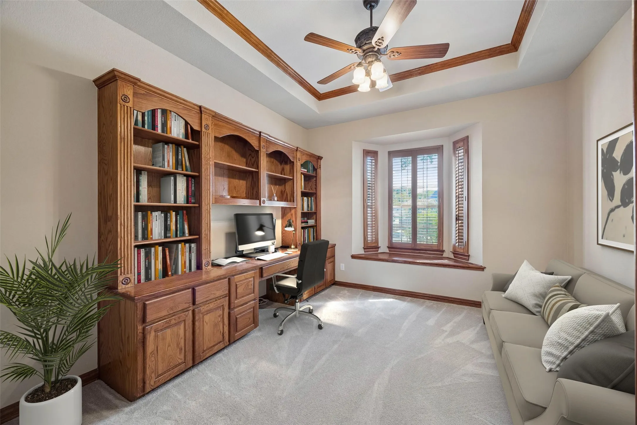 The study is found just off the entry and offers built-in desk with shelves and storage, tray ceiling with crown molding and ceiling fan, window seat with plantation shutters, carpet and a storage closet. (Virtually staged to show potential.)