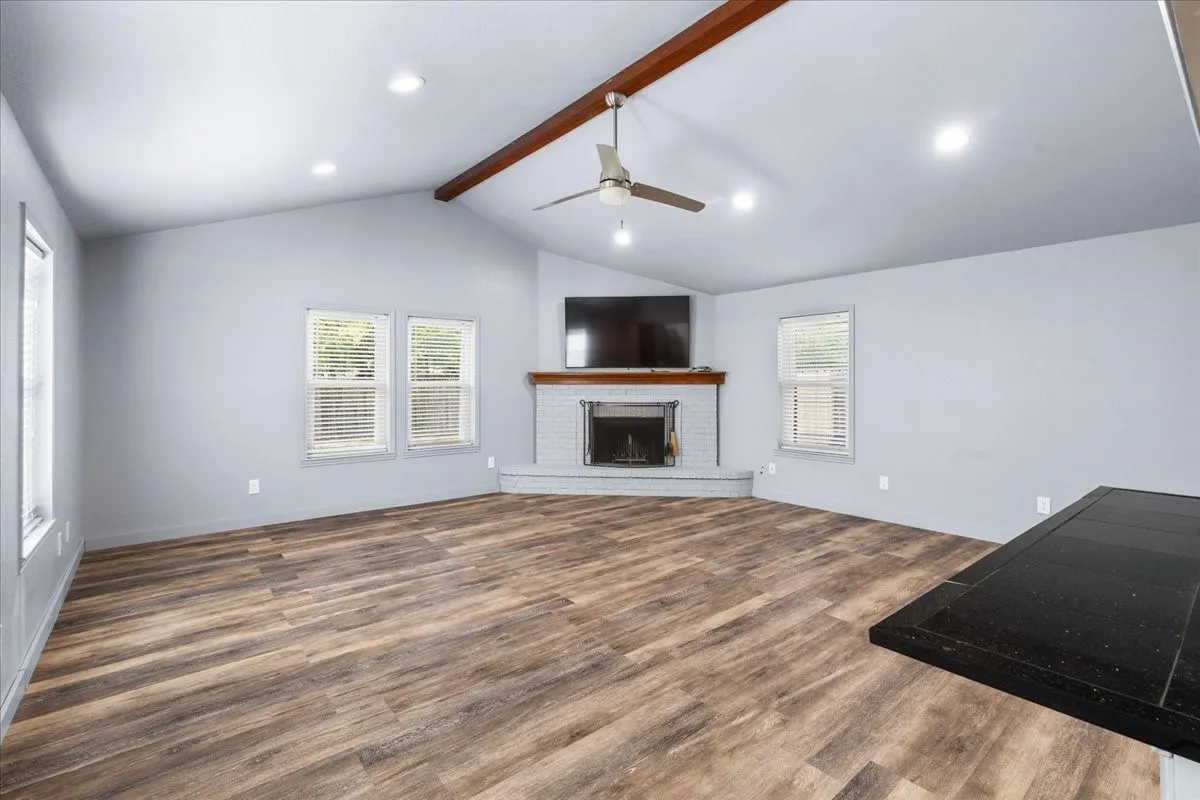 Vaulted ceiling with exposed wood beam, wood-finish flooring, recessed lighting, and a corner fireplace with a white brick surround