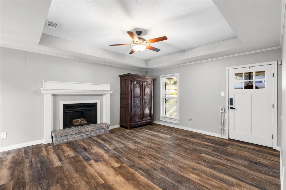 Wood-finish flooring, tray ceiling, ceiling fan, white door with glass panels, and a fireplace with a brick hearth