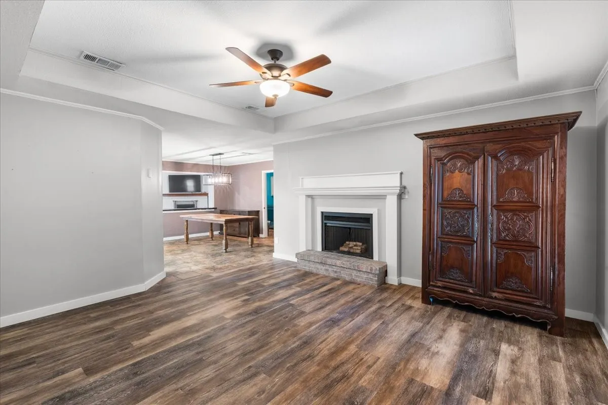 Tray ceiling with an attached ceiling fan, wood-finish flooring, and a fireplace with a white mantel and hearth