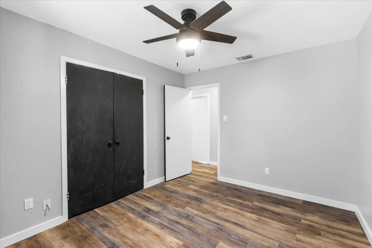 Wood-finish flooring, light gray walls, black bi-fold closet doors, white trim, and a ceiling fan with integrated lighting