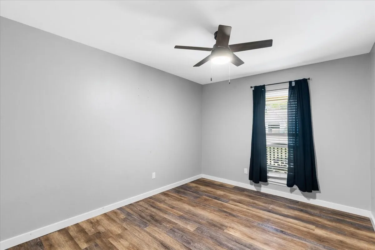 Wood-finish flooring, light gray walls, window with horizontal blinds, and a ceiling fan with integrated lighting
