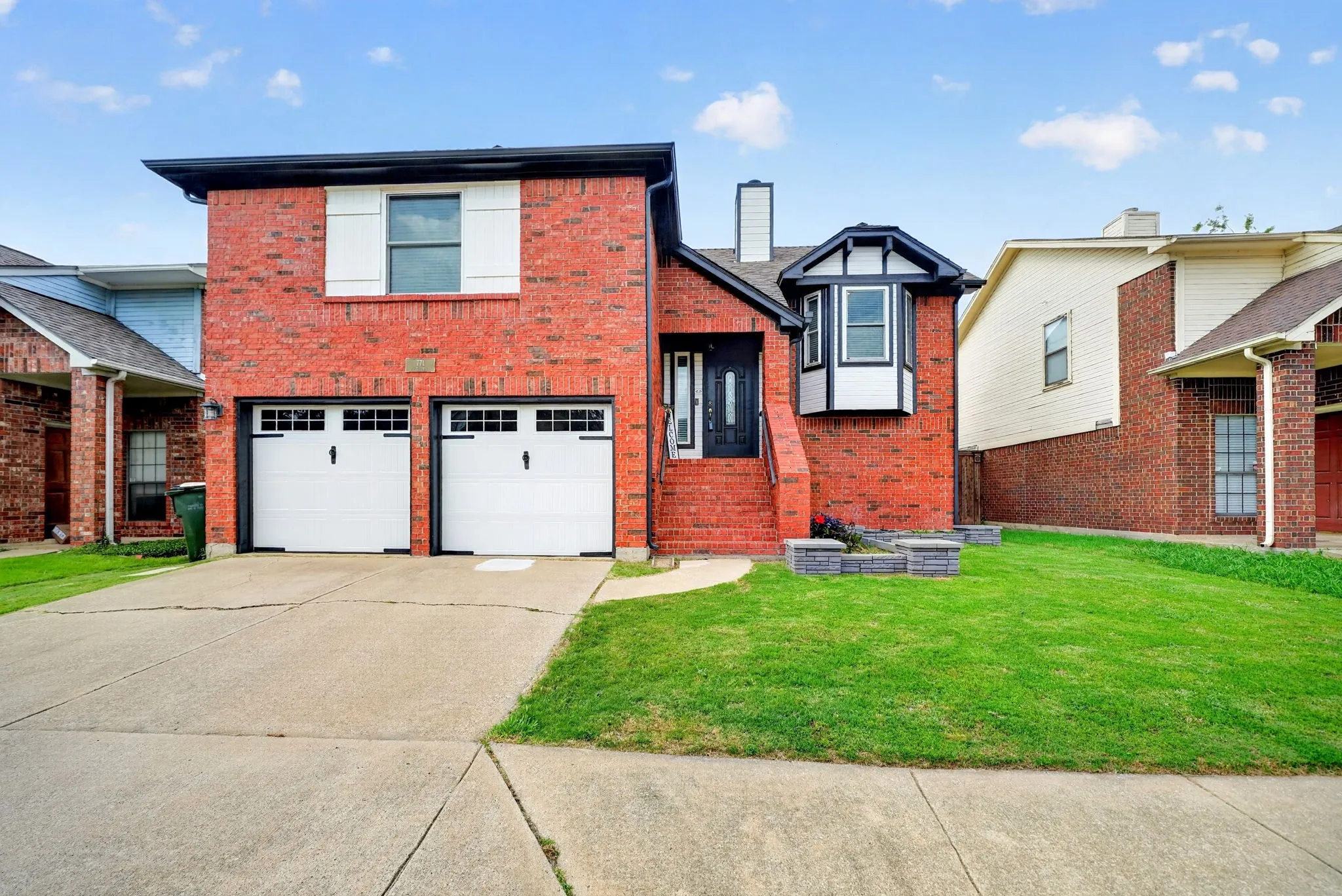 Brick exterior with white window shutters, two white garage doors, a concrete driveway, and a manicured lawn