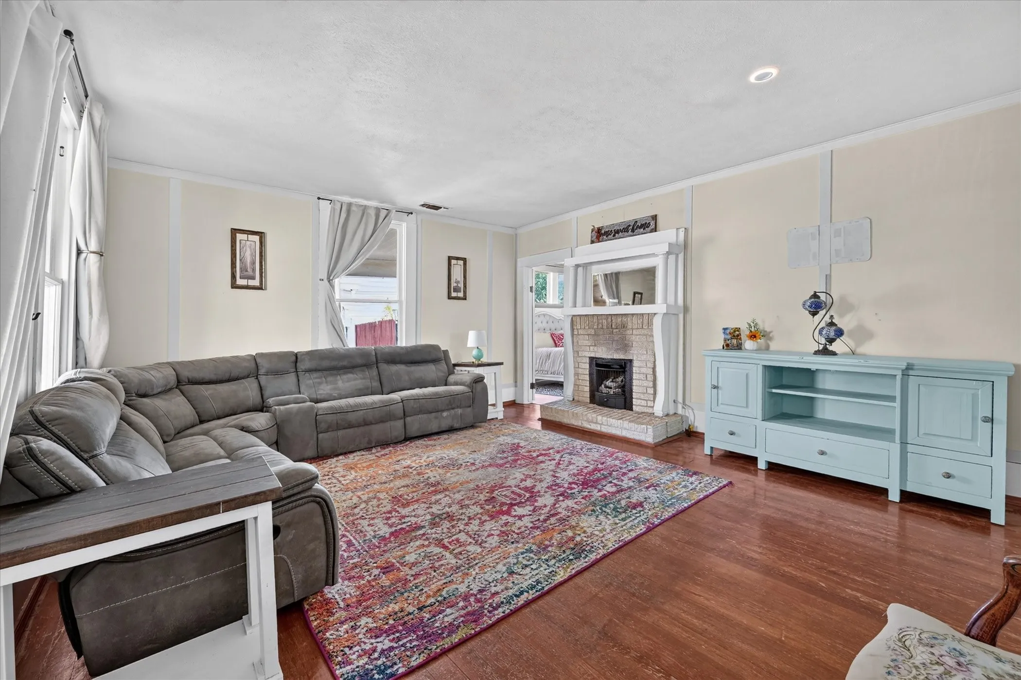 Hardwood flooring, brick fireplace with decorative mantel, recessed ceiling light, multiple windows, and a built-in media console