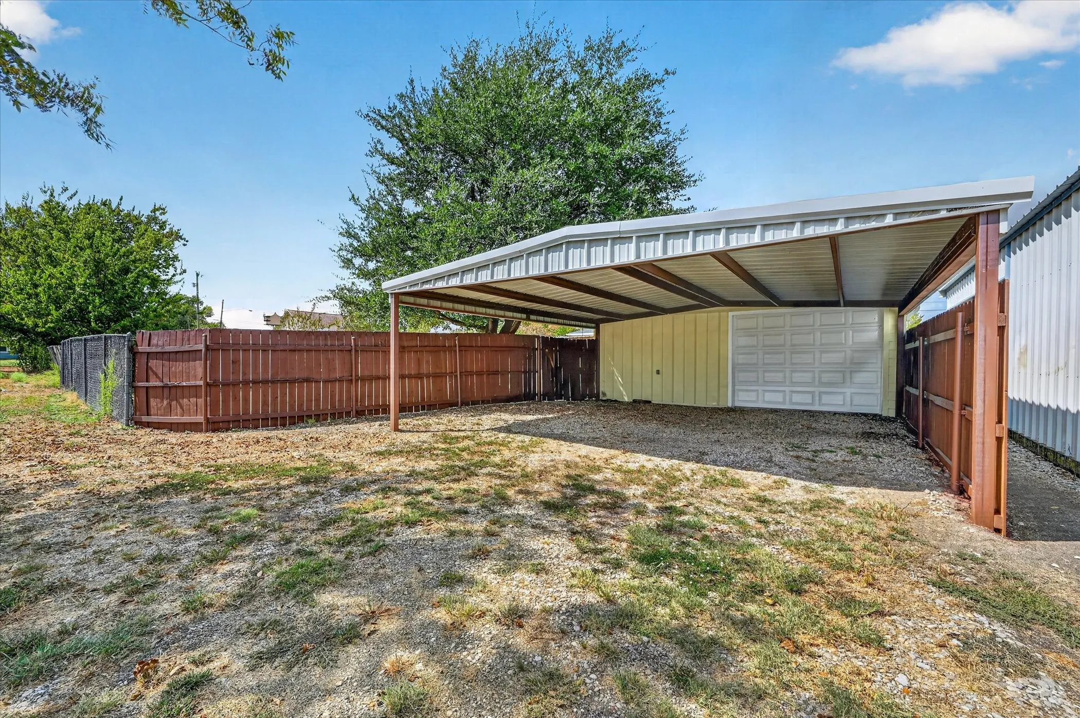 Carport with metal roof and support beams, privacy fencing, and gravel parking surface