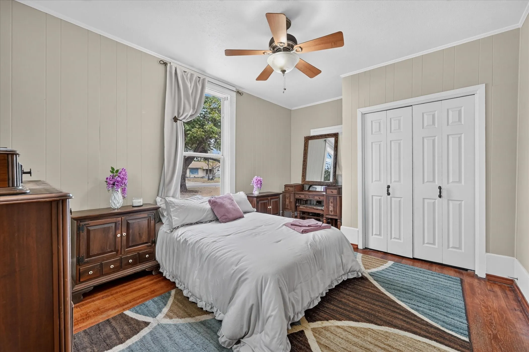 Hardwood-style flooring, paneling wall treatment, ceiling fan with light fixture, window with drapes, and bi-fold closet doors